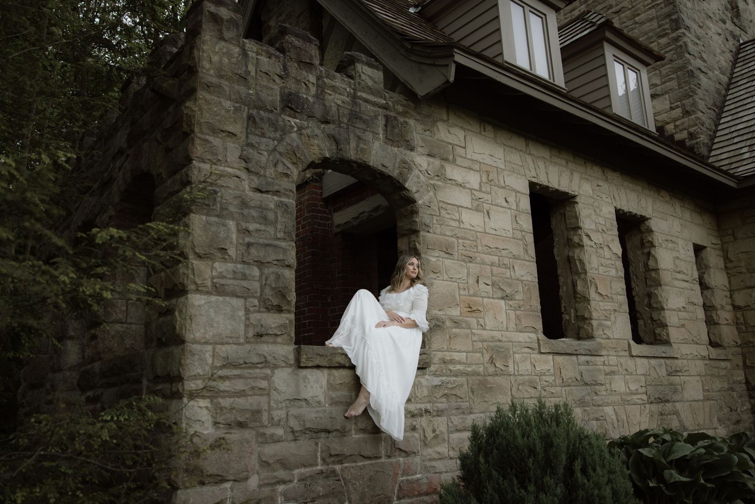 A figure in a white flowy robe leans elegantly through a stone window opening of a historic building.