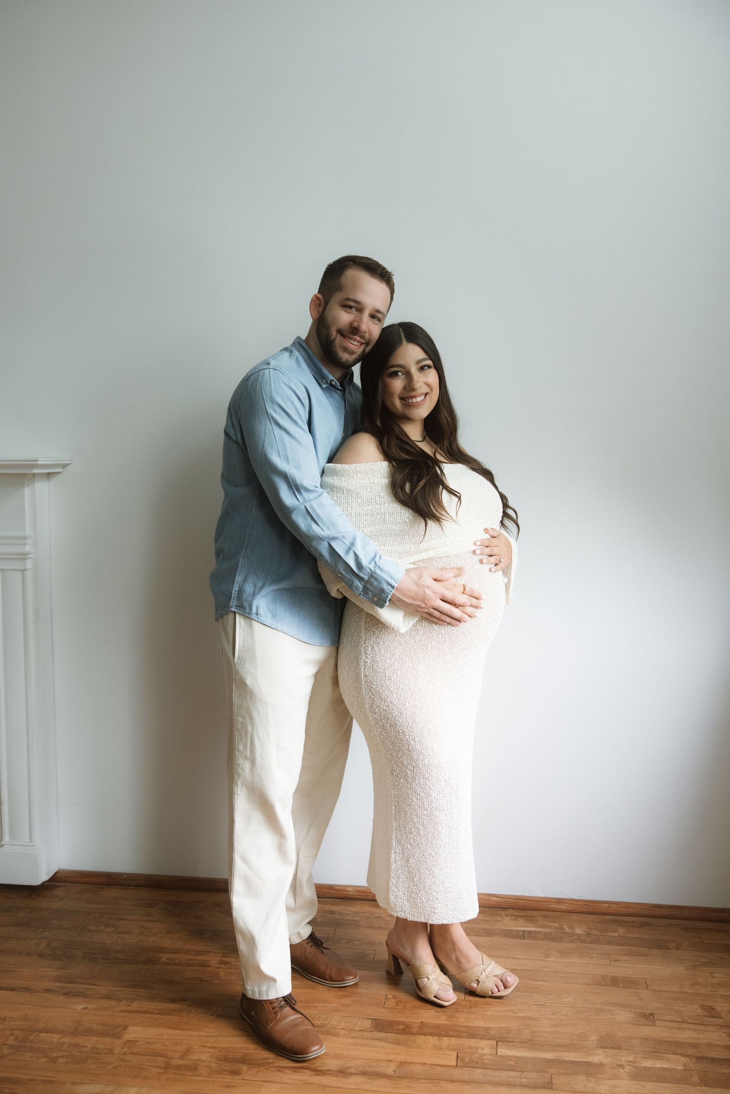 Romantic photo session shows couple embracing on wooden floor against minimalist white wall with natural light.
