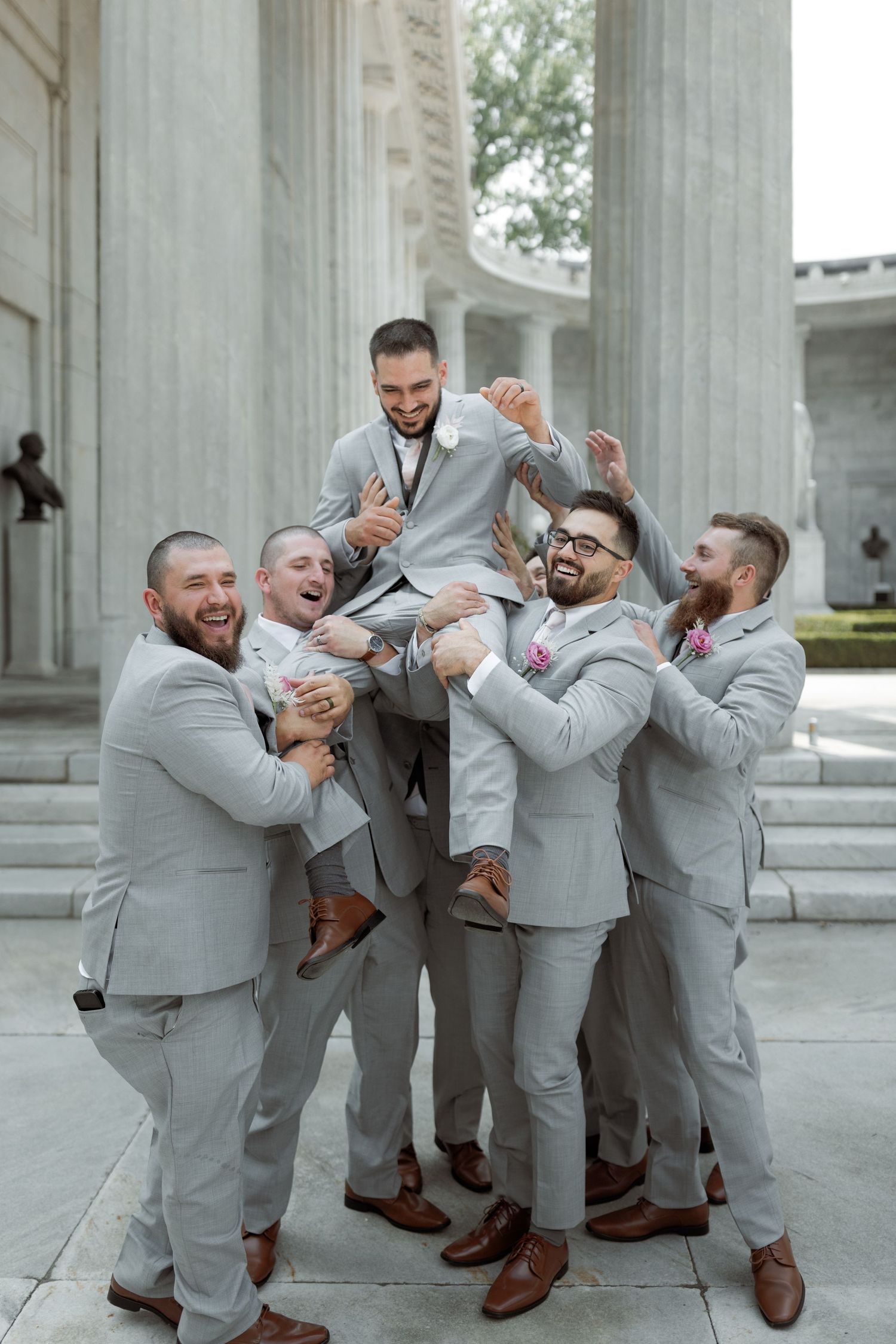 Groomsmen in light gray suits playfully lift and celebrate with the groom outside a classical columned building.
