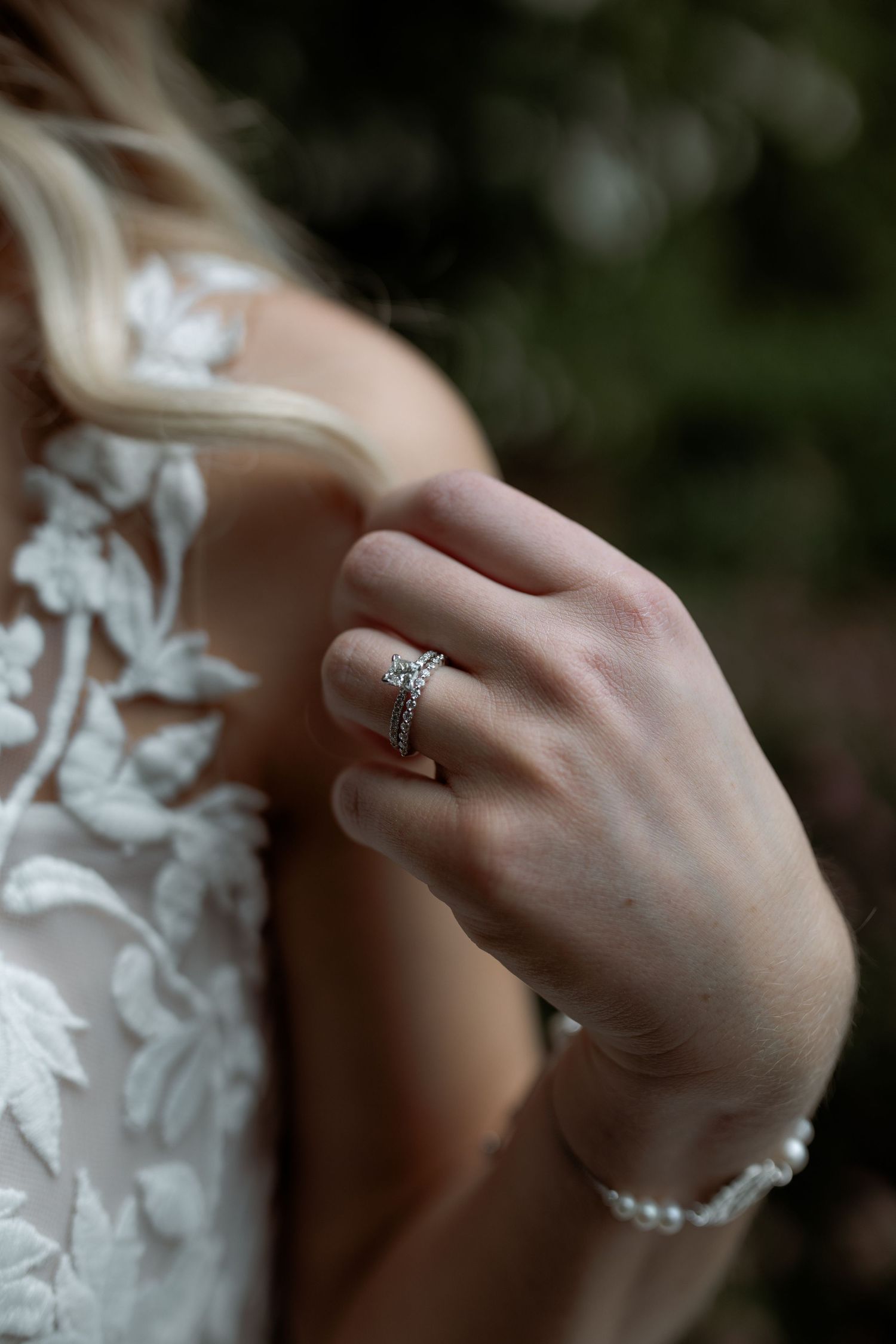 Close-up of a bride's hand wearing an elegant engagement ring and delicate bracelet with lace dress details.