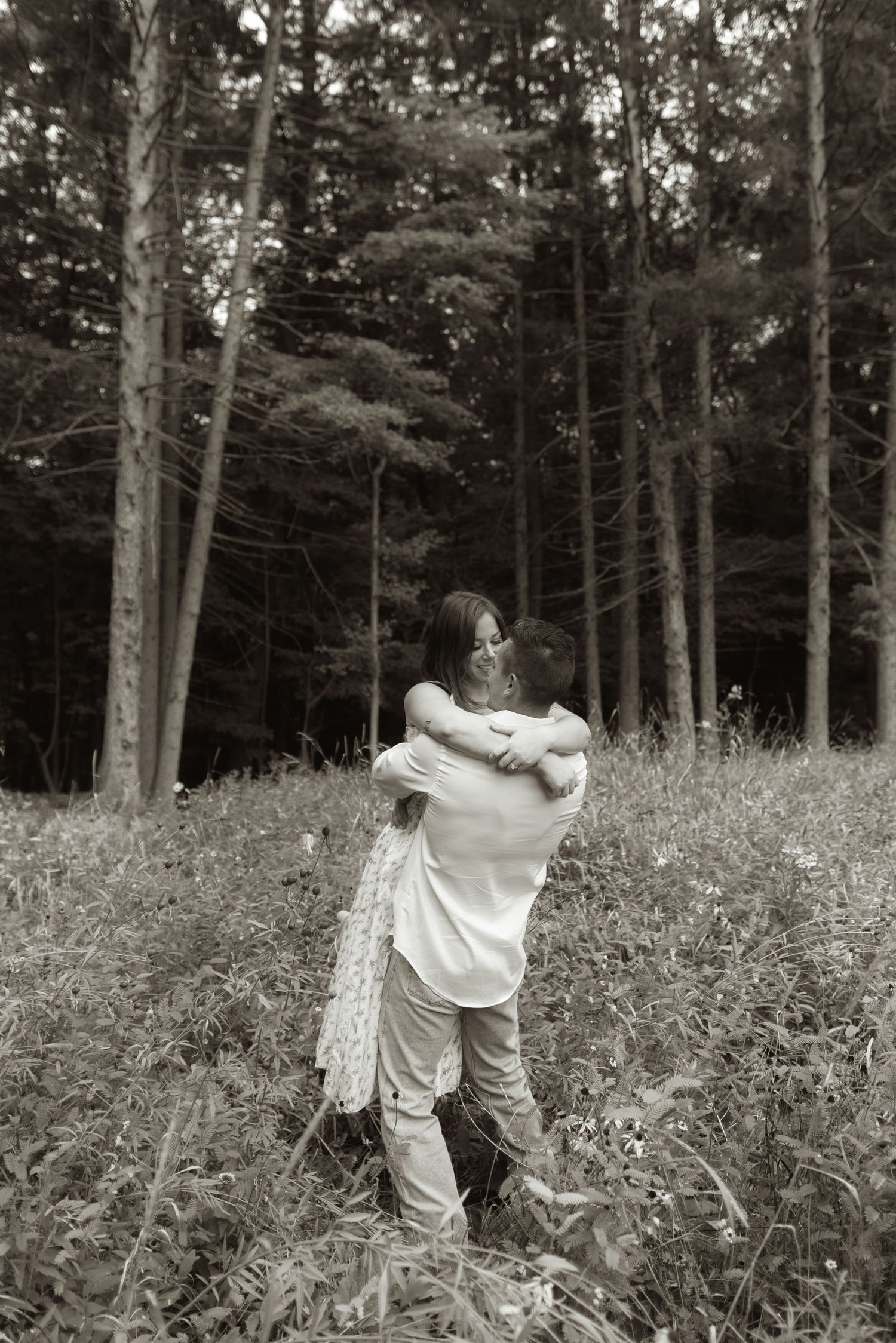 A romantic black and white photo of a couple embracing in a wooded forest clearing.