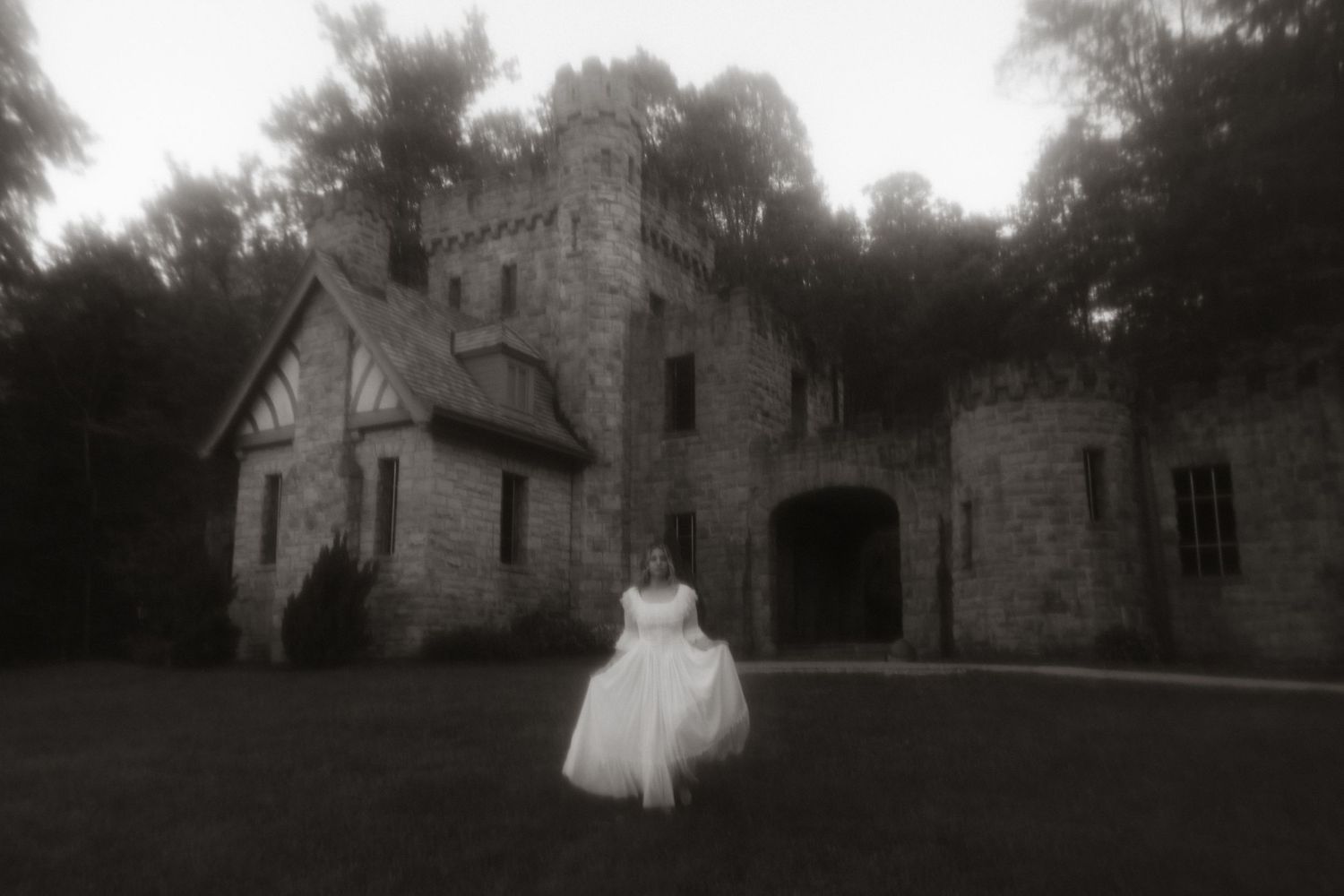 A figure in a white dress stands before a Gothic-style stone mansion in this ethereal black and white photograph.
