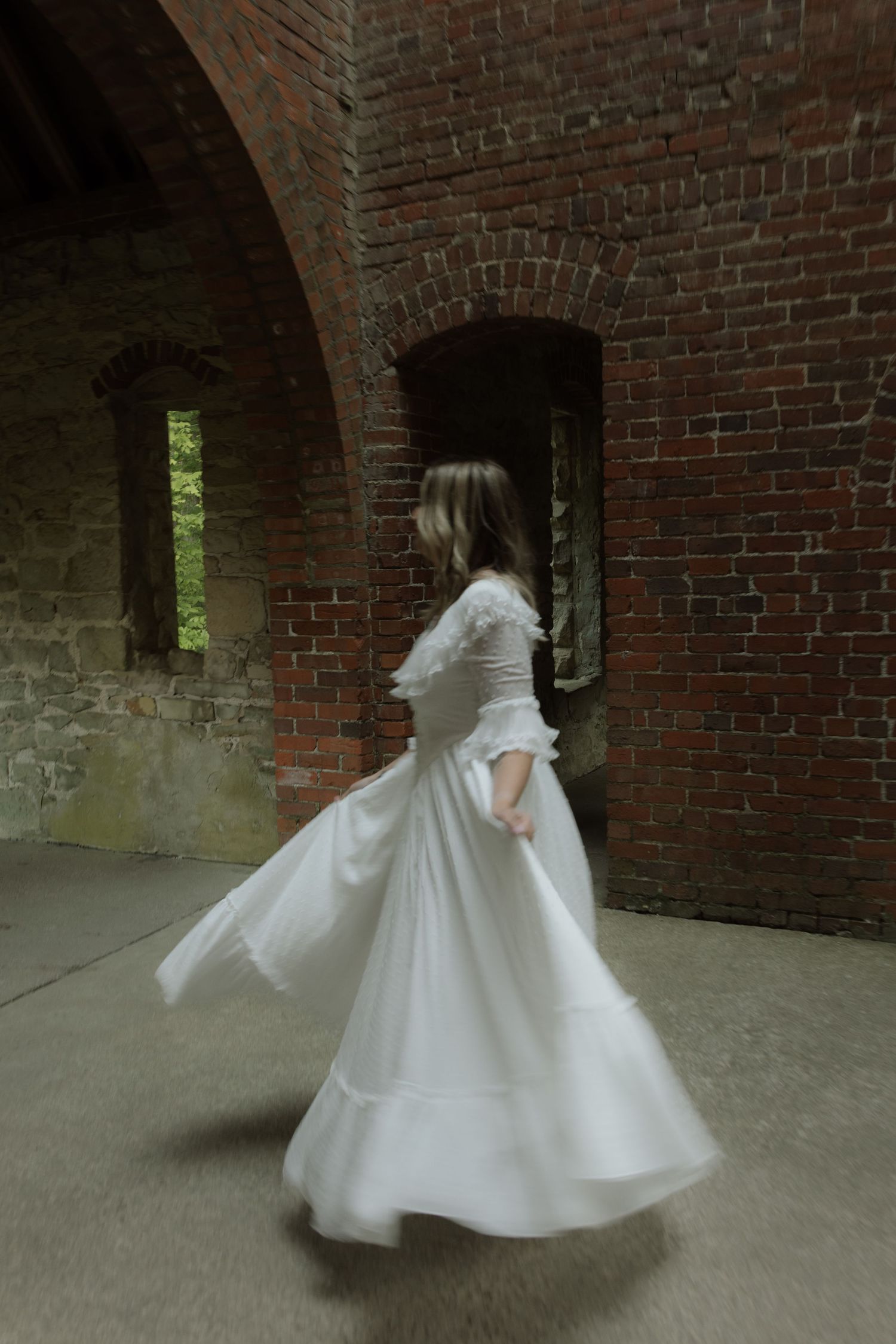 A figure in a flowing white vintage dress twirls inside a brick archway with natural light filtering through.