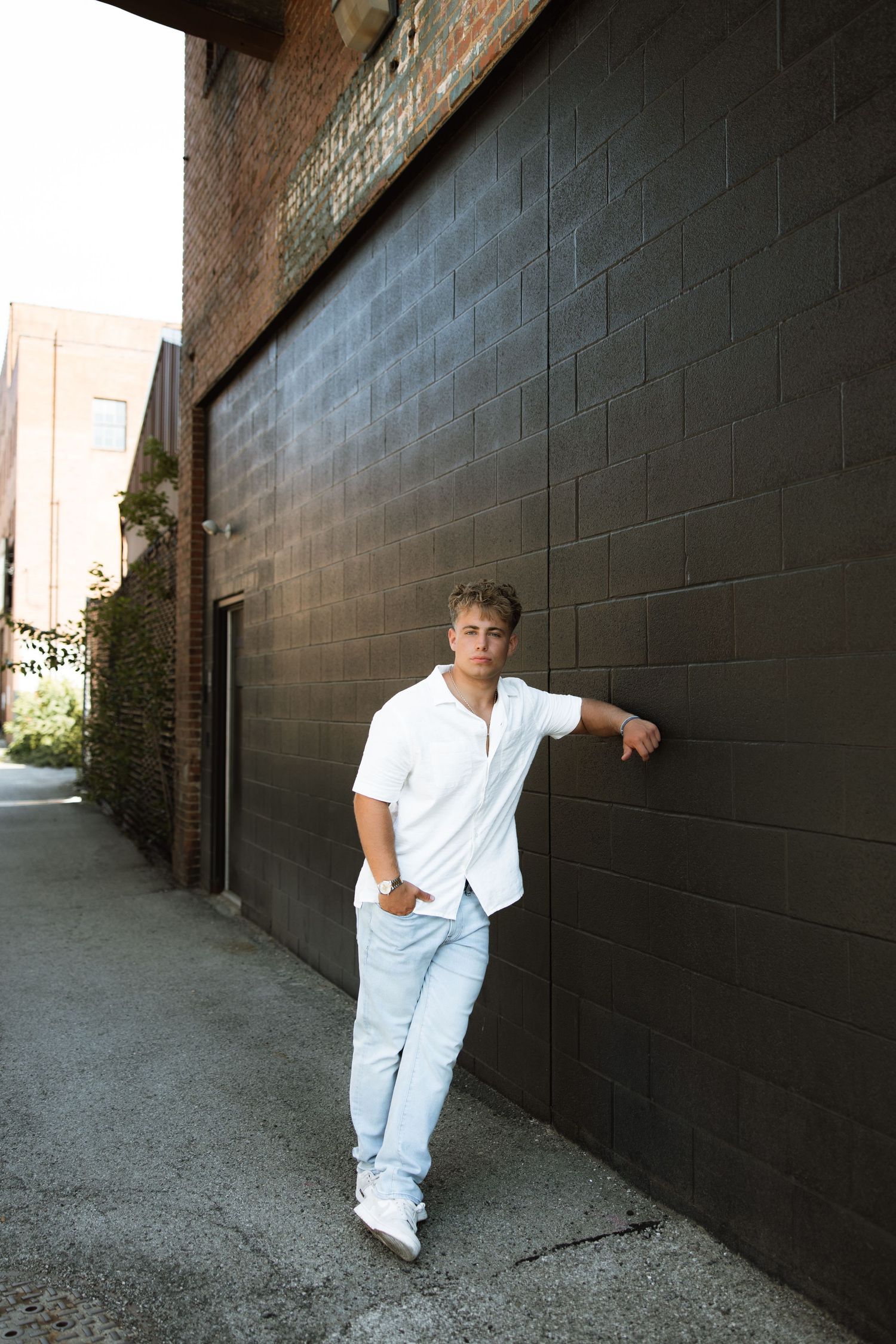 Person in white shirt and light jeans leaning against black brick wall in urban alleyway setting.