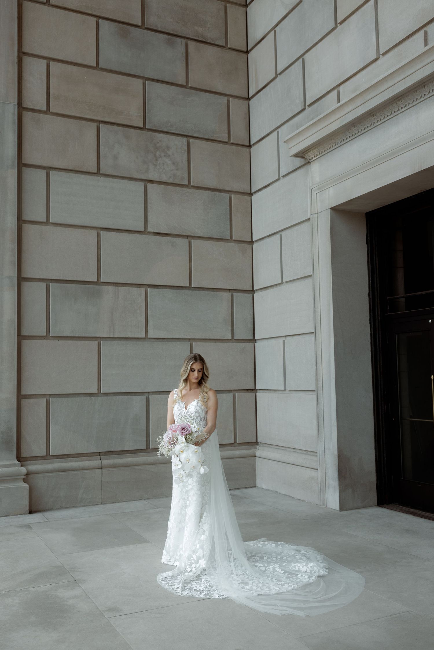 A bride in a white lace wedding gown with long train poses elegantly against a stone wall near a doorway.
