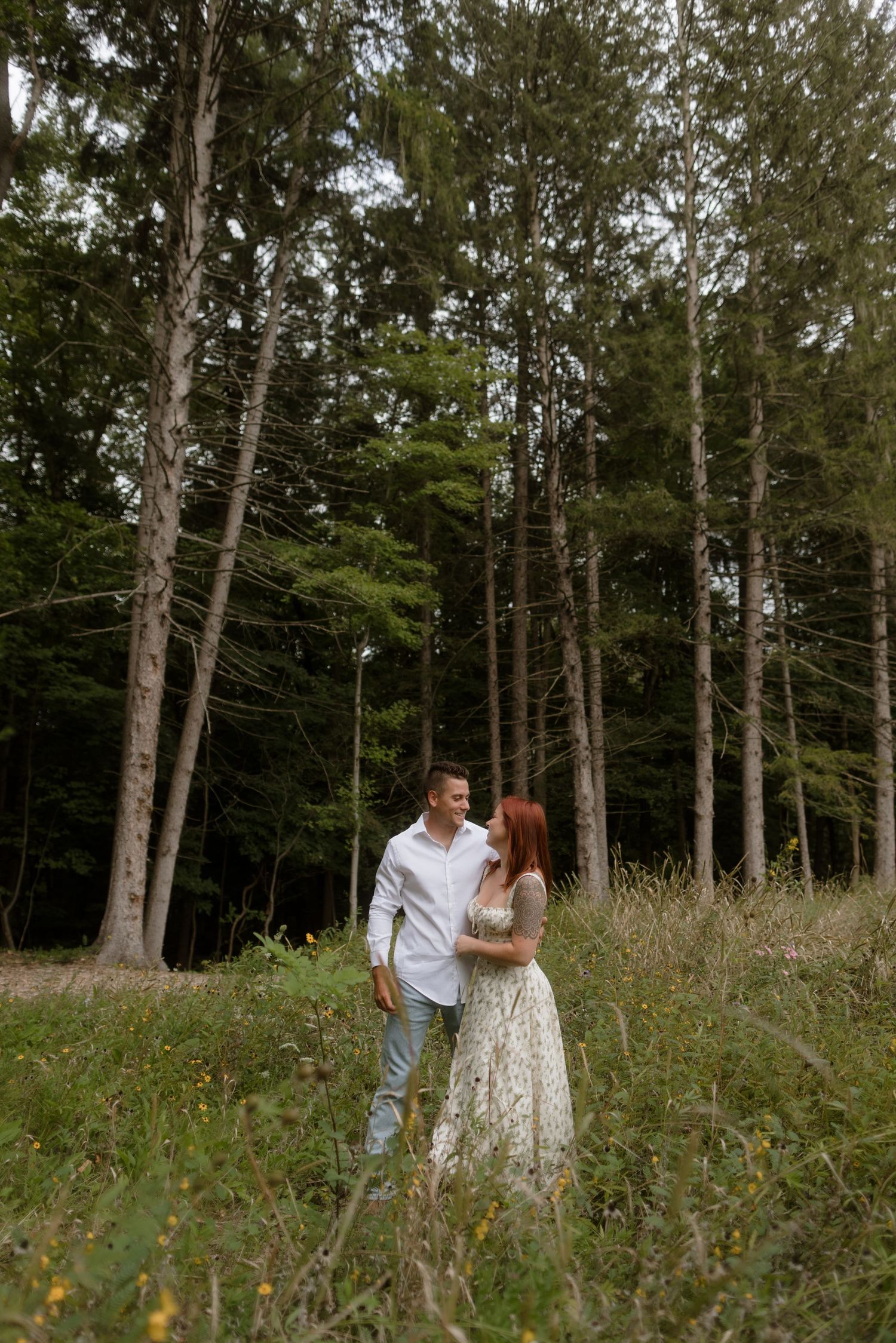 A romantic couple walks together in a sunlit forest, surrounded by tall trees and wild grass.