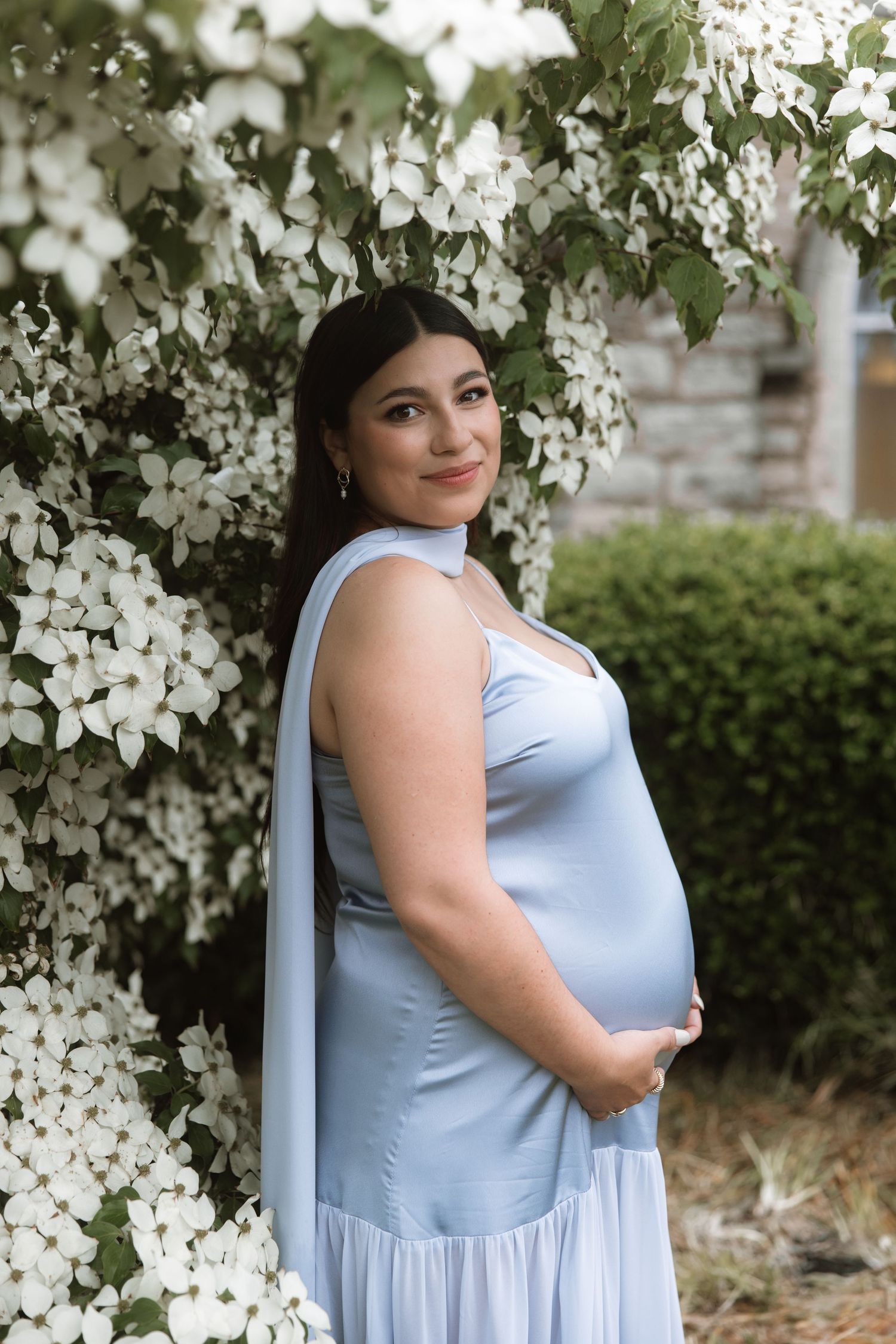 Maternity portrait with person in light blue dress among blooming white flowers.