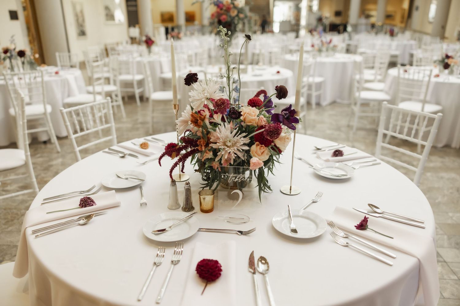 Elegant wedding reception table setting with a burgundy and white floral centerpiece on a round table with white linens.
