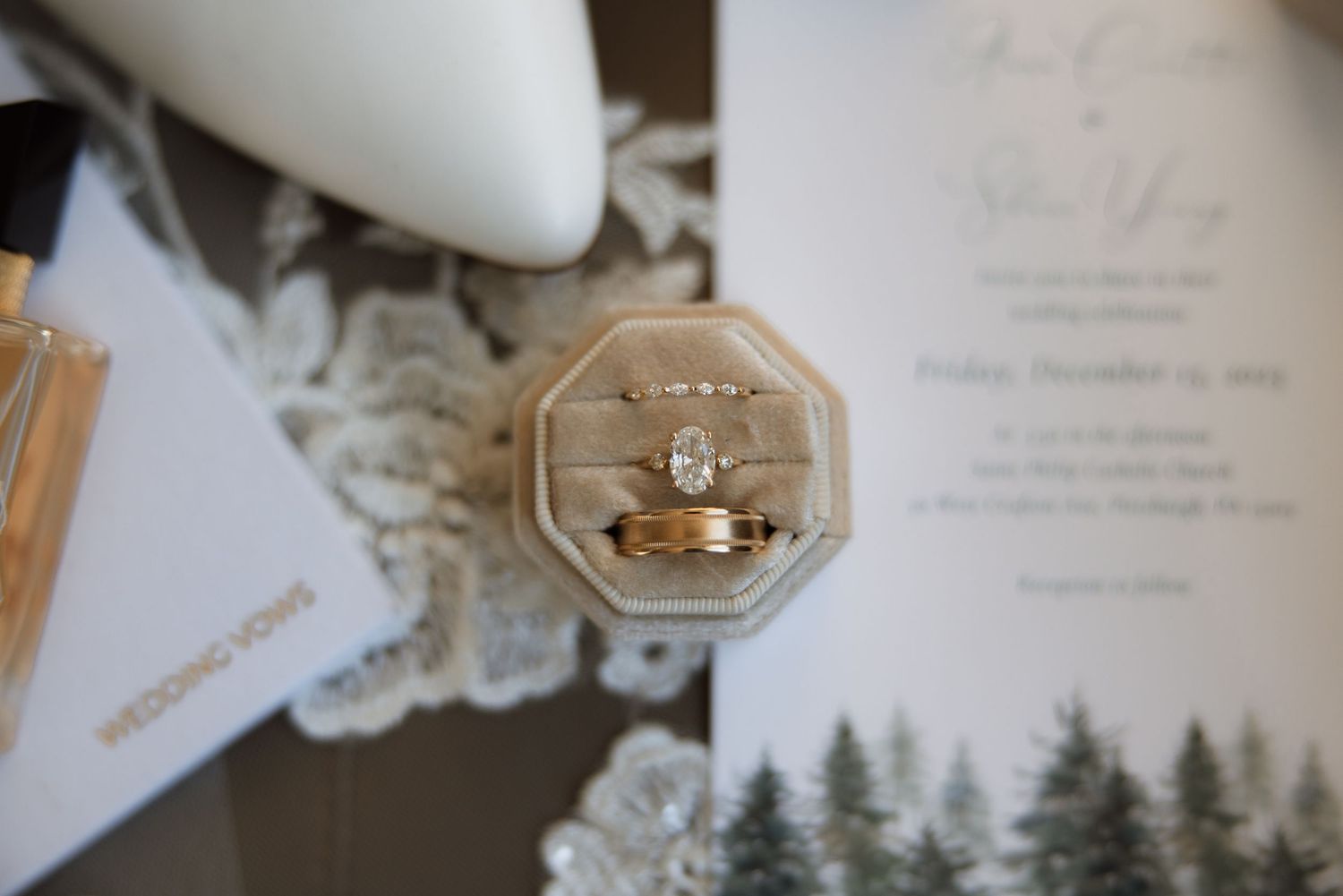 Gold wedding rings displayed in a hexagonal ring box with pine tree silhouettes and lace decor in the background.