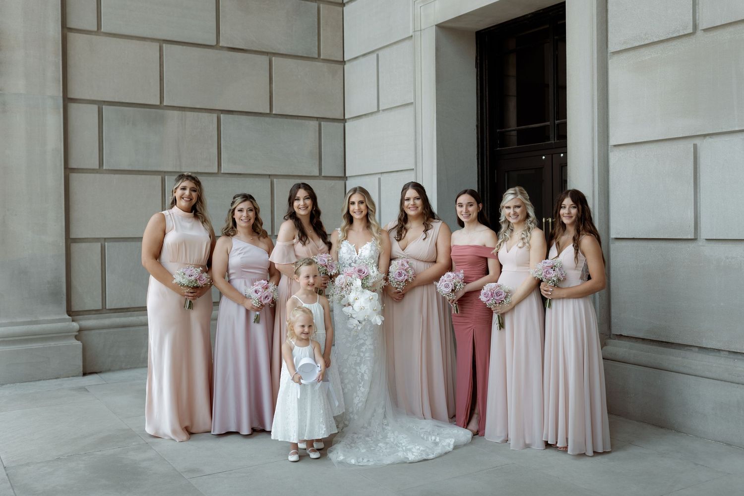 Bridesmaids in flowing blush and pink dresses pose with flower girl and bride in white lace gown against stone wall.