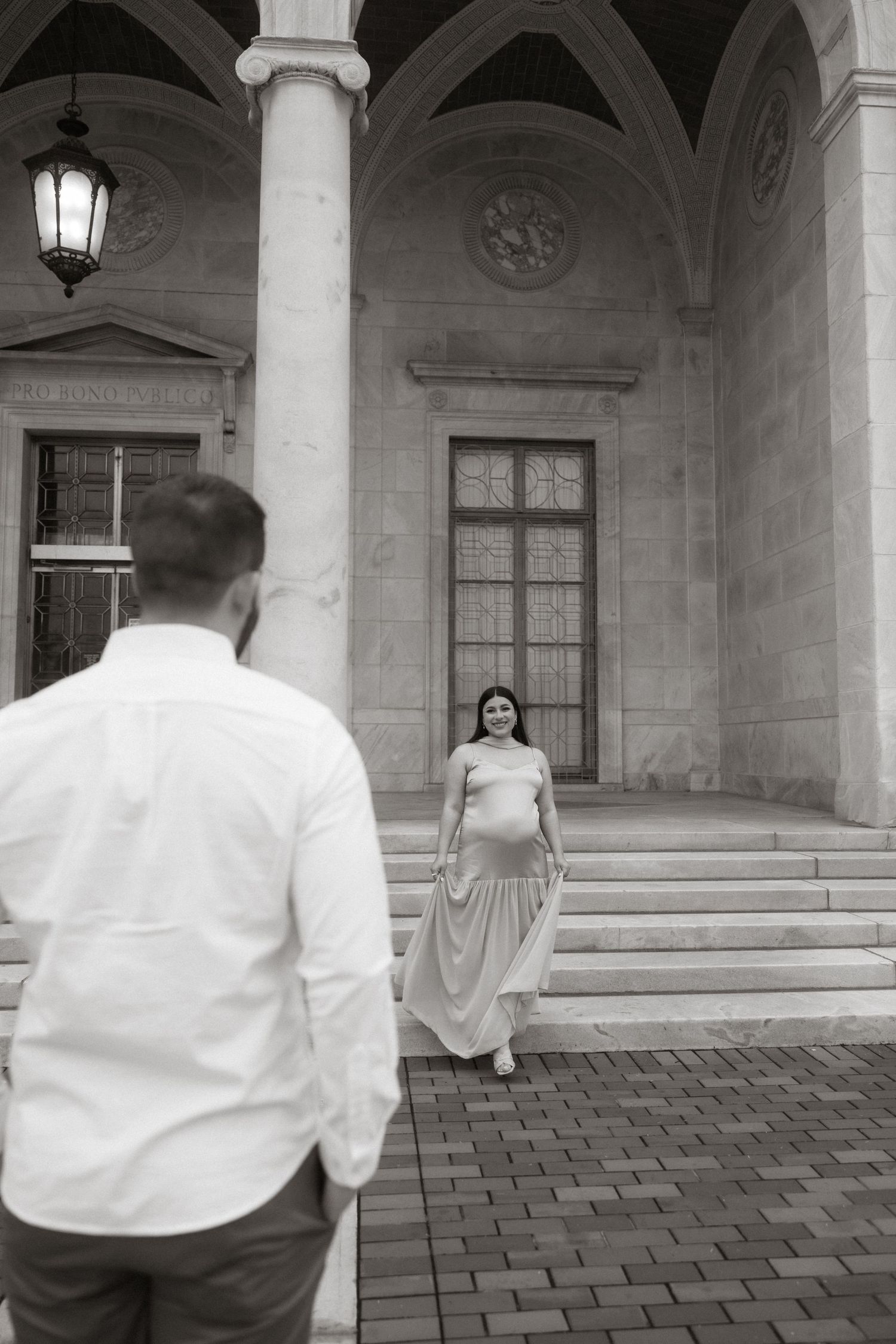 A black and white photo showing a couple posing on steps of an elegant building with arched architecture.