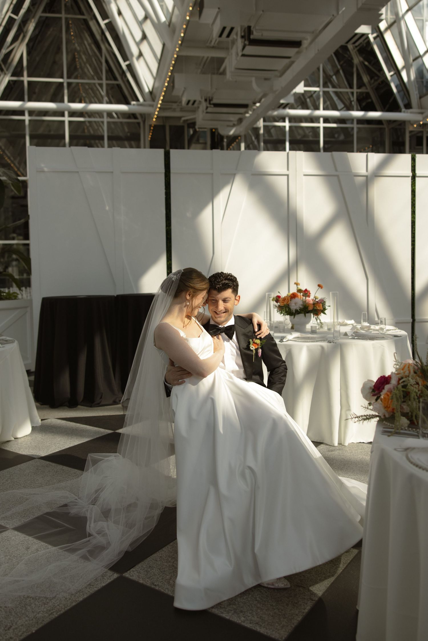 Bride and groom share an intimate moment in a sunlit conservatory with decorative table settings.
