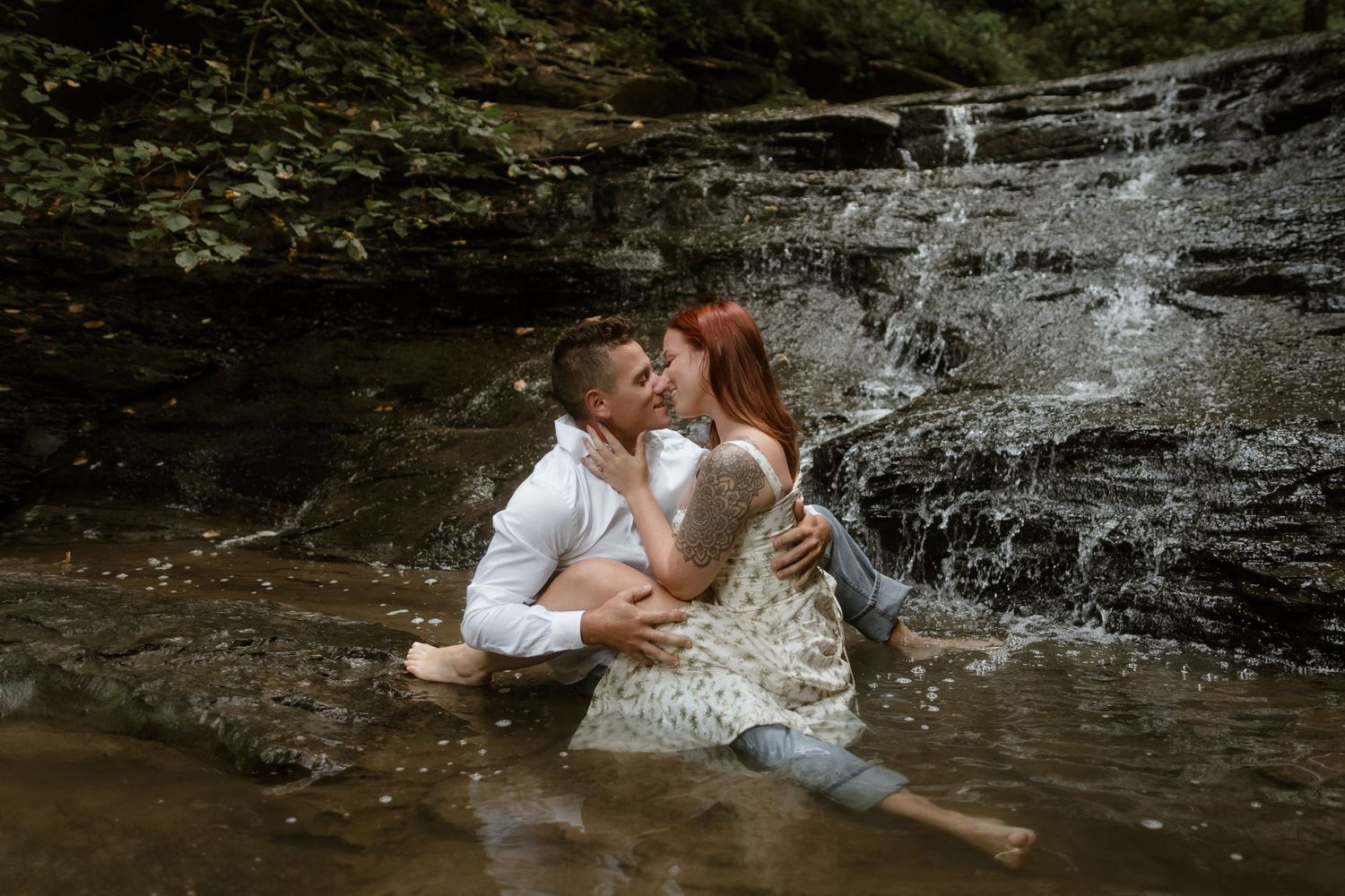 A couple shares a romantic moment while sitting in shallow water at the base of a scenic waterfall.