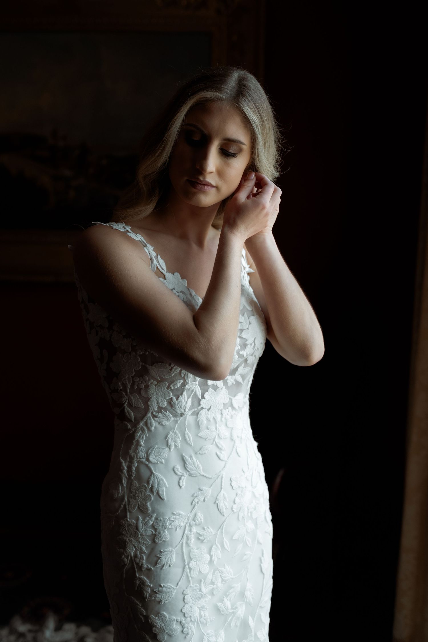 A bride in an elegant white lace wedding dress poses in moody lighting against a dark background.