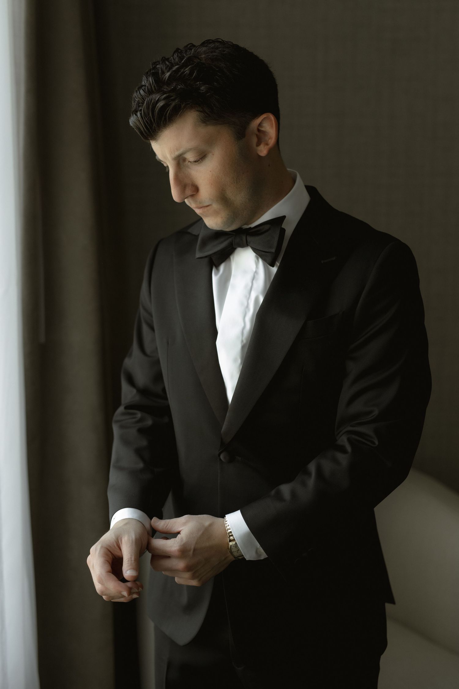 Groom in classic black tuxedo adjusting cuffs by window light.