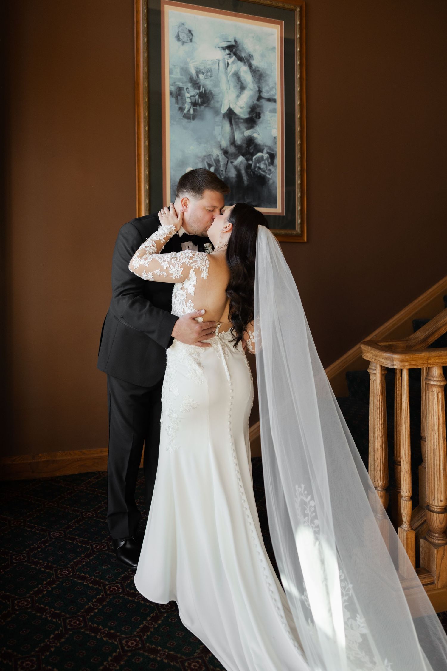A couple in wedding attire share romantic moments on a staircase with vintage photographs on the wall behind them.