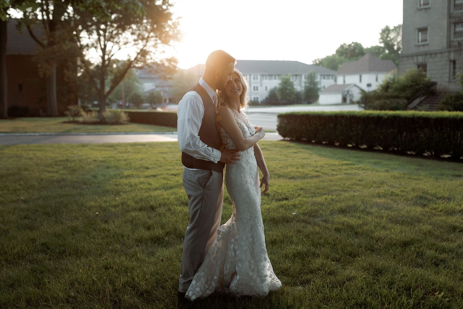 Couple shares intimate moment at sunset on grassy lawn in wedding attire.