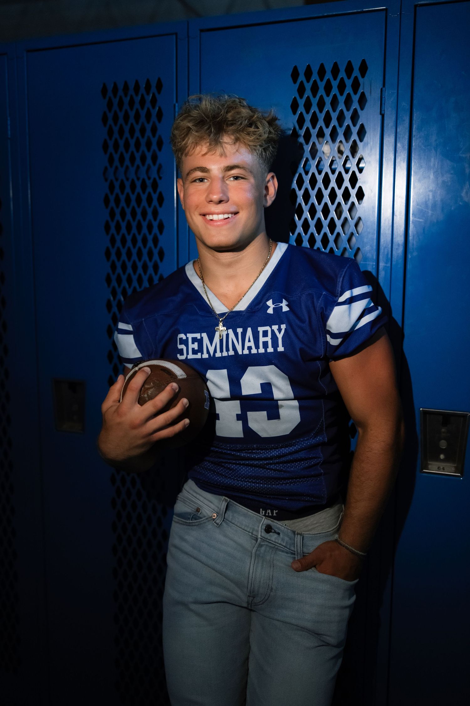 A student athlete in blue Seminary jersey #13 poses with a football by blue lockers.