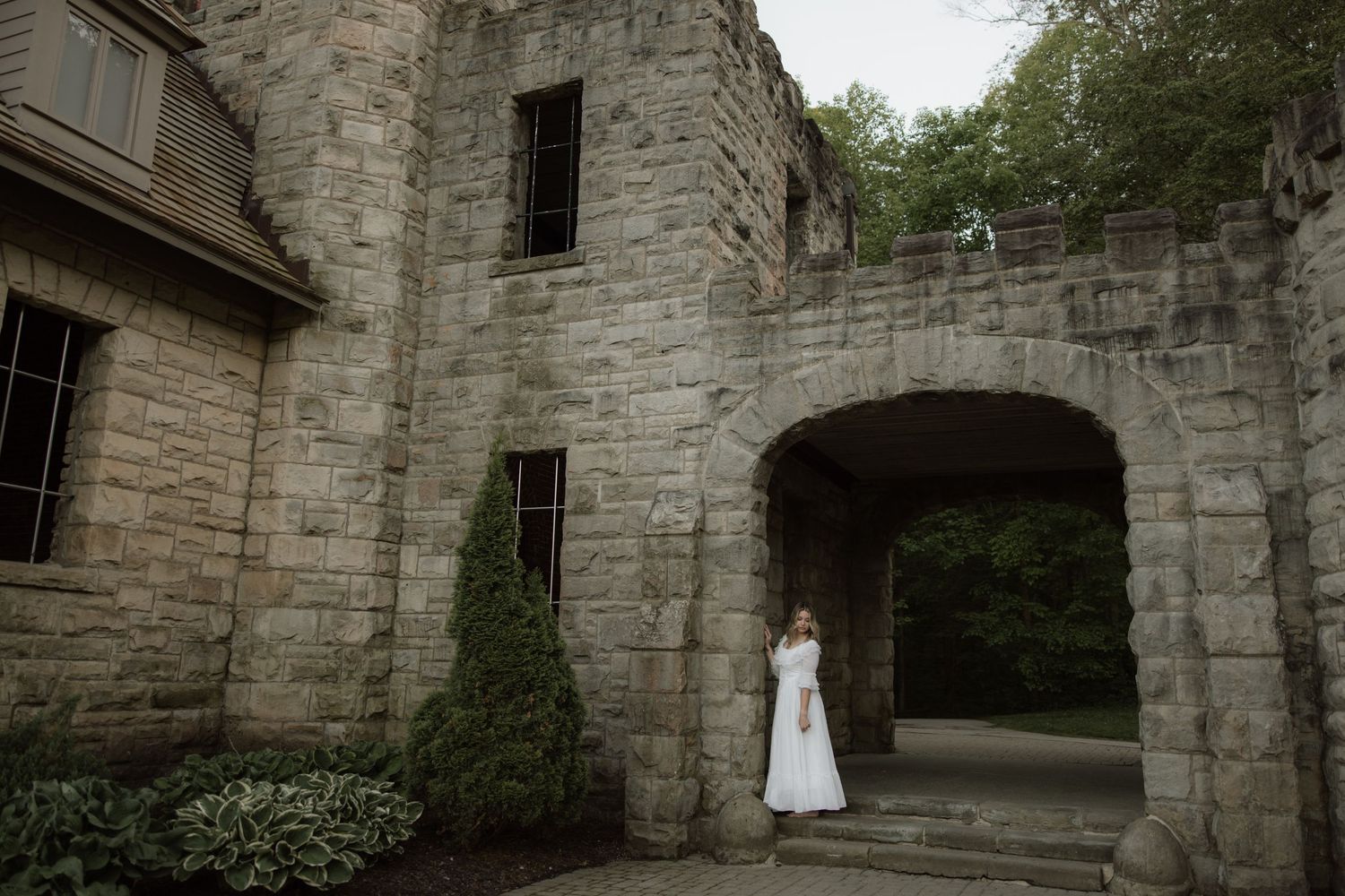 A figure in a white dress stands under a stone archway entrance of a historic castle-like building surrounded by greenery.
