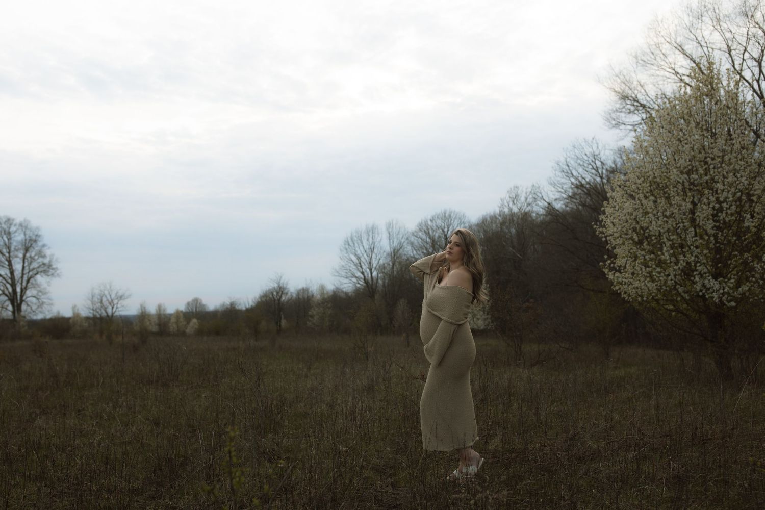 Person in beige dress stands alone in a misty field with bare trees in background.