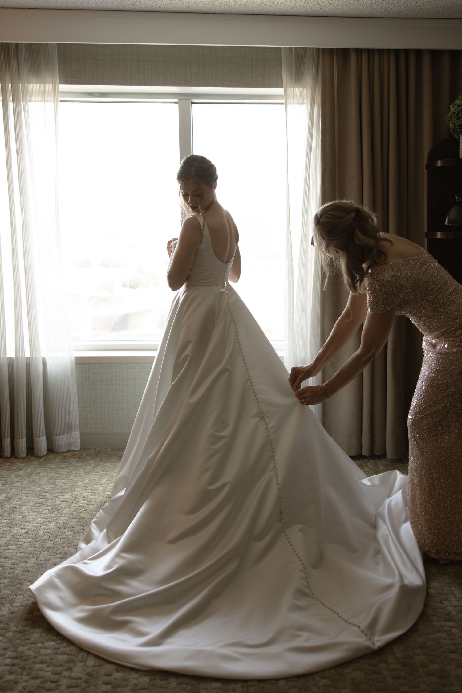 Helper adjusting long train of white wedding gown in sunlit hotel room.