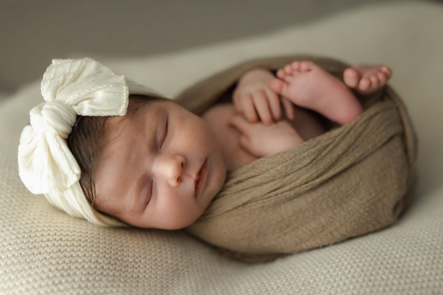 Newborn baby swaddled in brown fabric with white bow headband sleeping peacefully on textured backdrop.