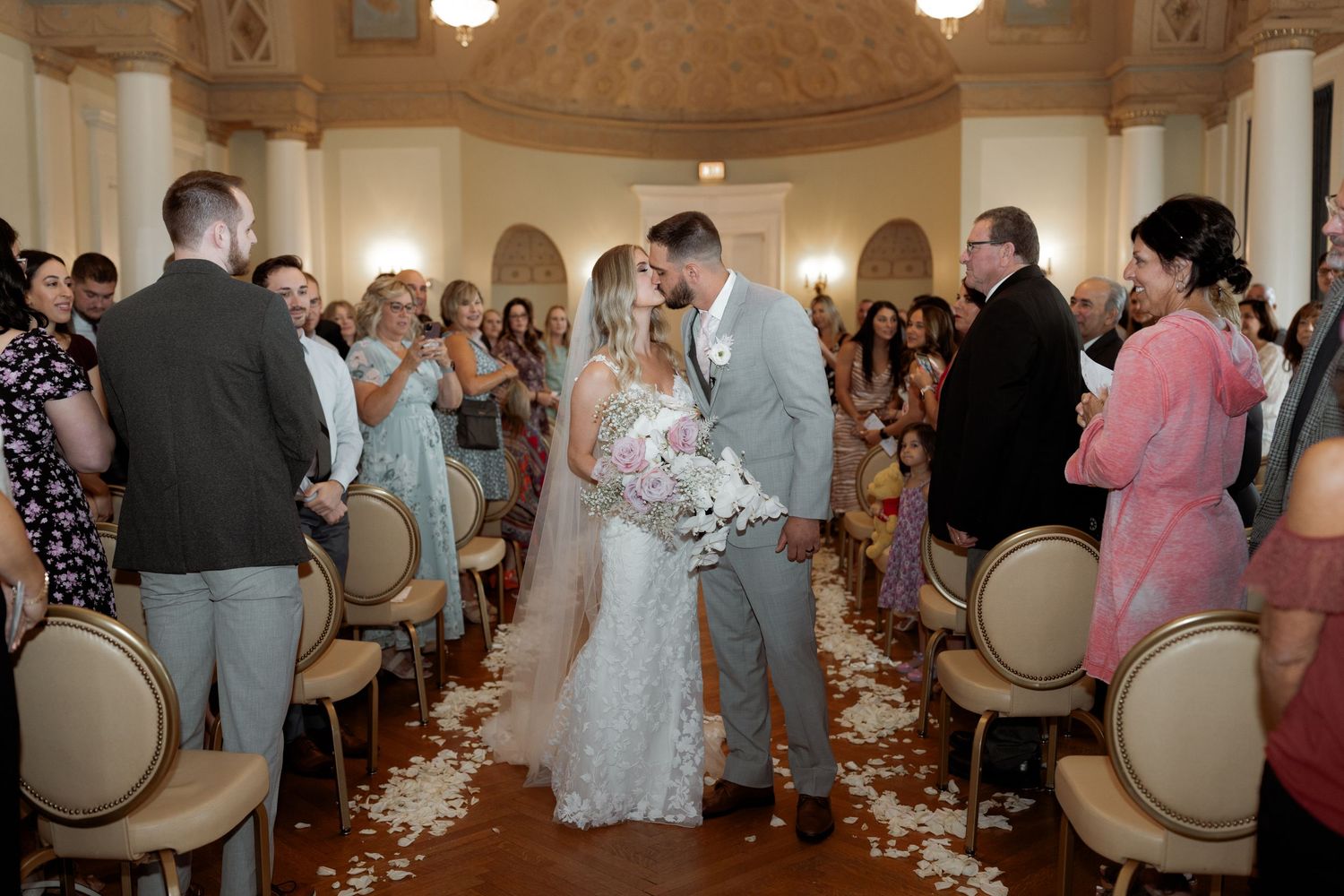 Newlyweds share their first kiss as guests celebrate in an elegant ballroom with rose gold chairs and scattered petals.