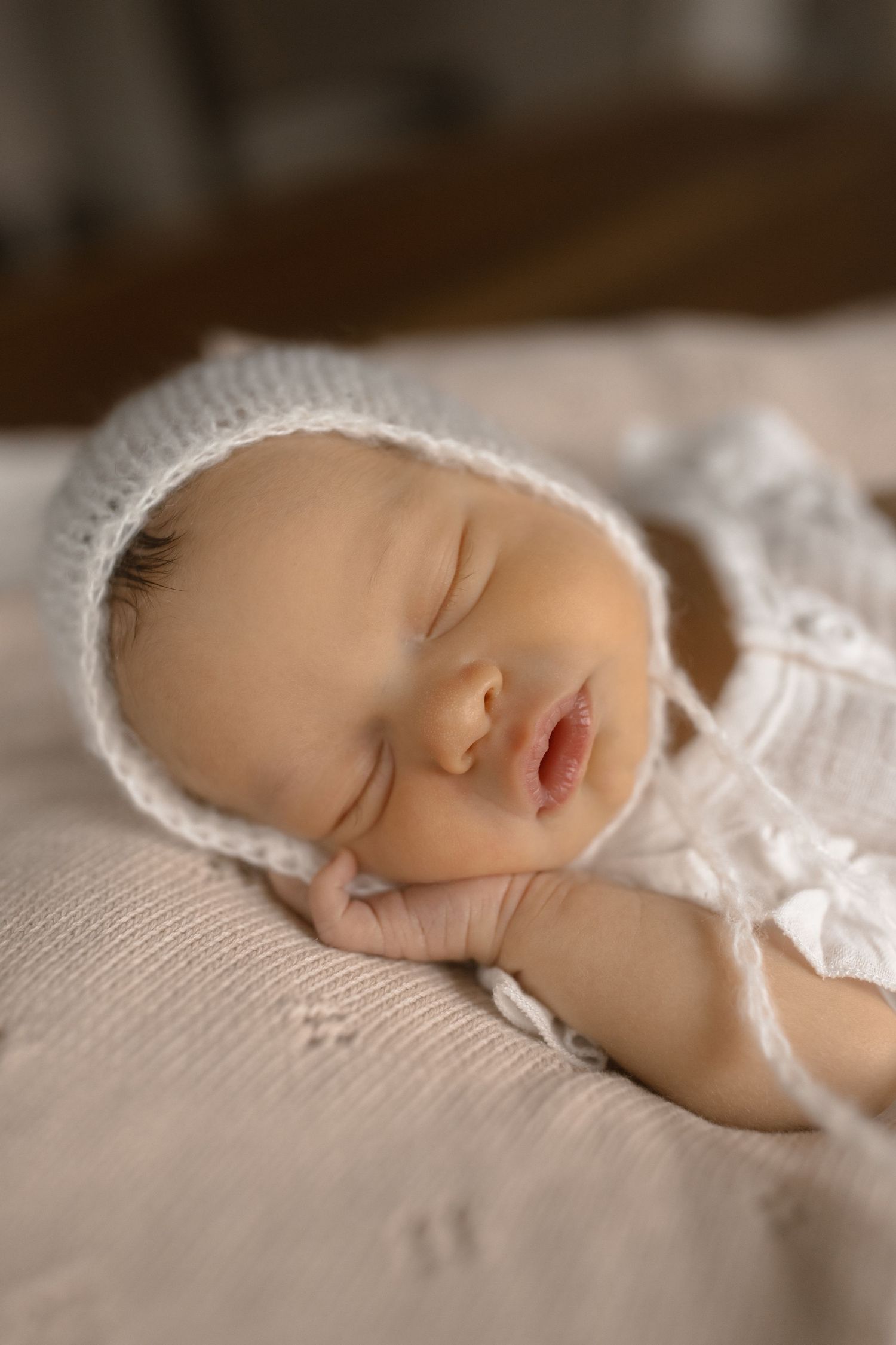 Peaceful sleeping newborn in white bonnet and knit clothing rests on textured fabric in soft lighting.