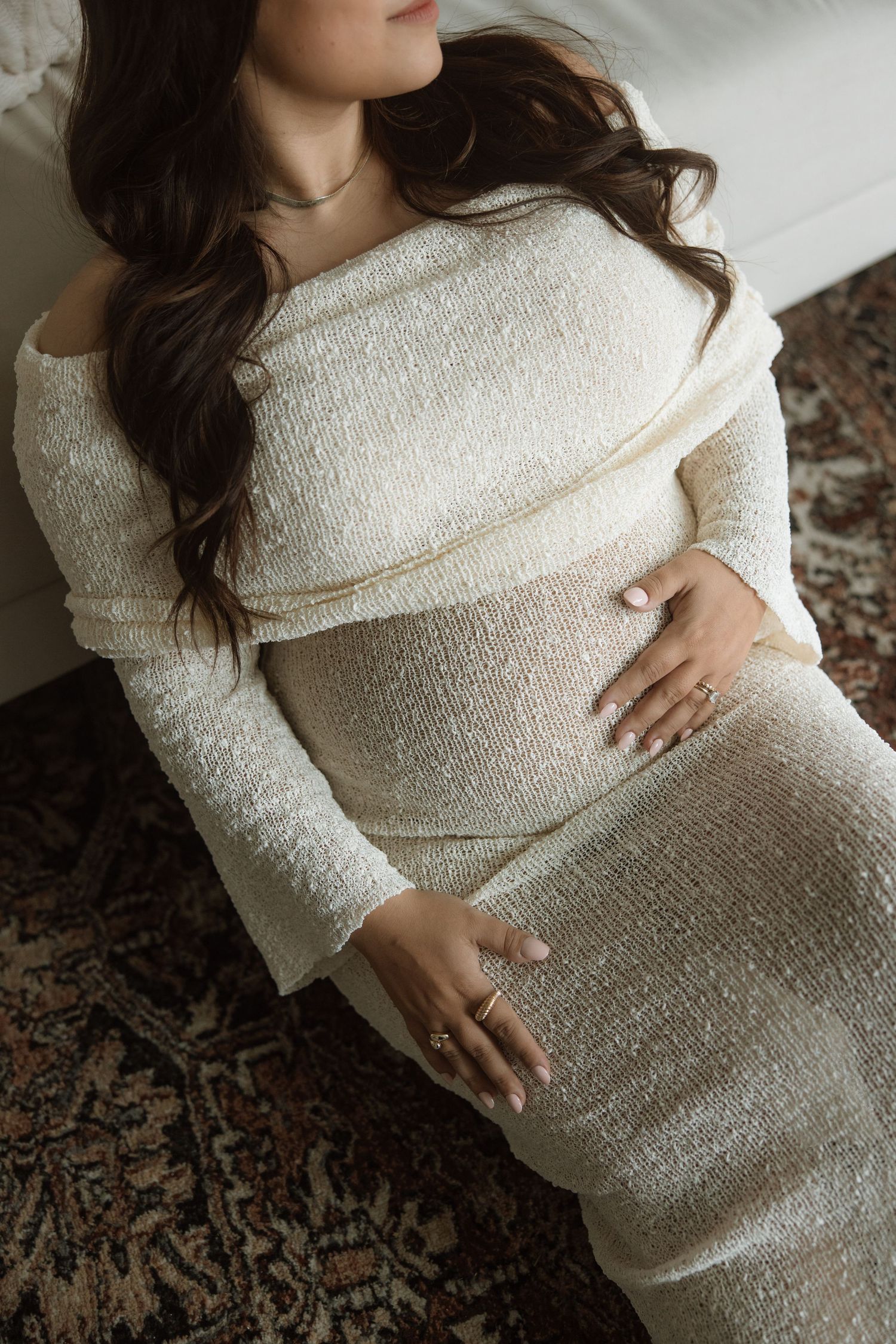 A pregnant person in a cream-colored knit dress rests their hand on their belly in a maternity photoshoot.