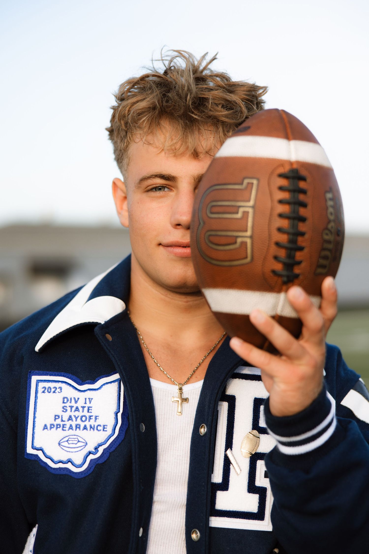 Student athlete in varsity jacket poses with football showing playoff state championship patch.