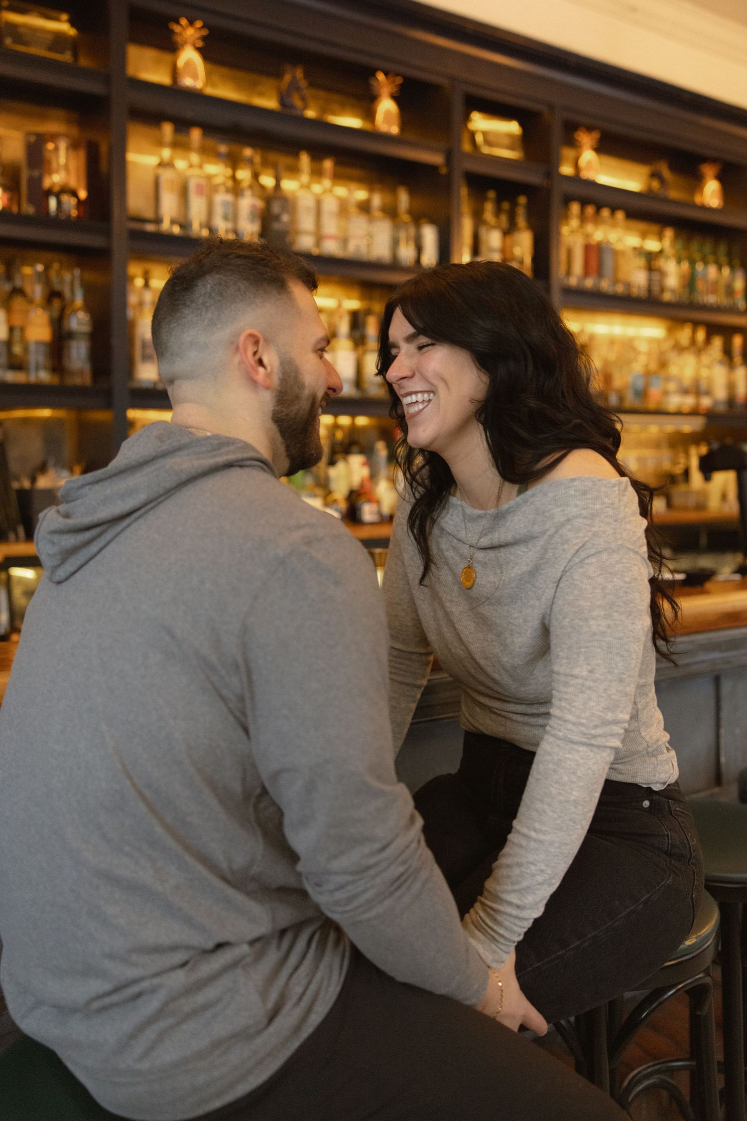 A couple shares a joyful moment while sitting at a bar with illuminated bottles in the background.