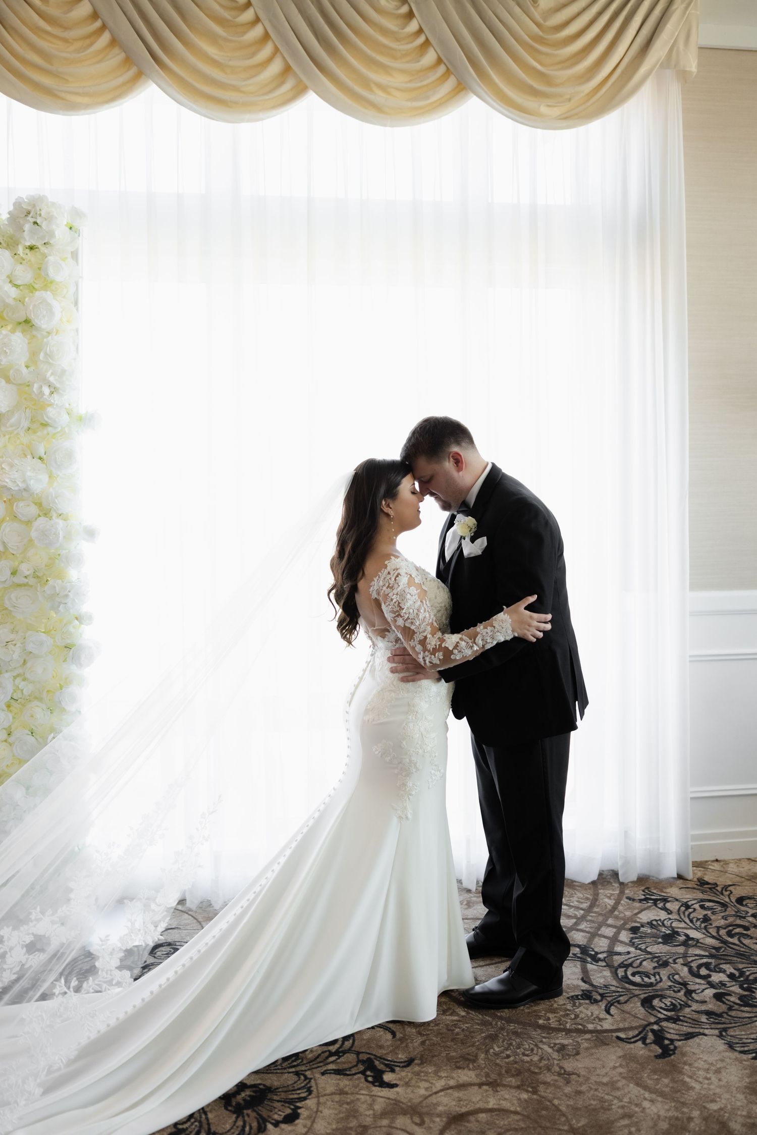 Elegant bride in lace sleeve wedding dress and groom share intimate moment by draped window with floral decor.