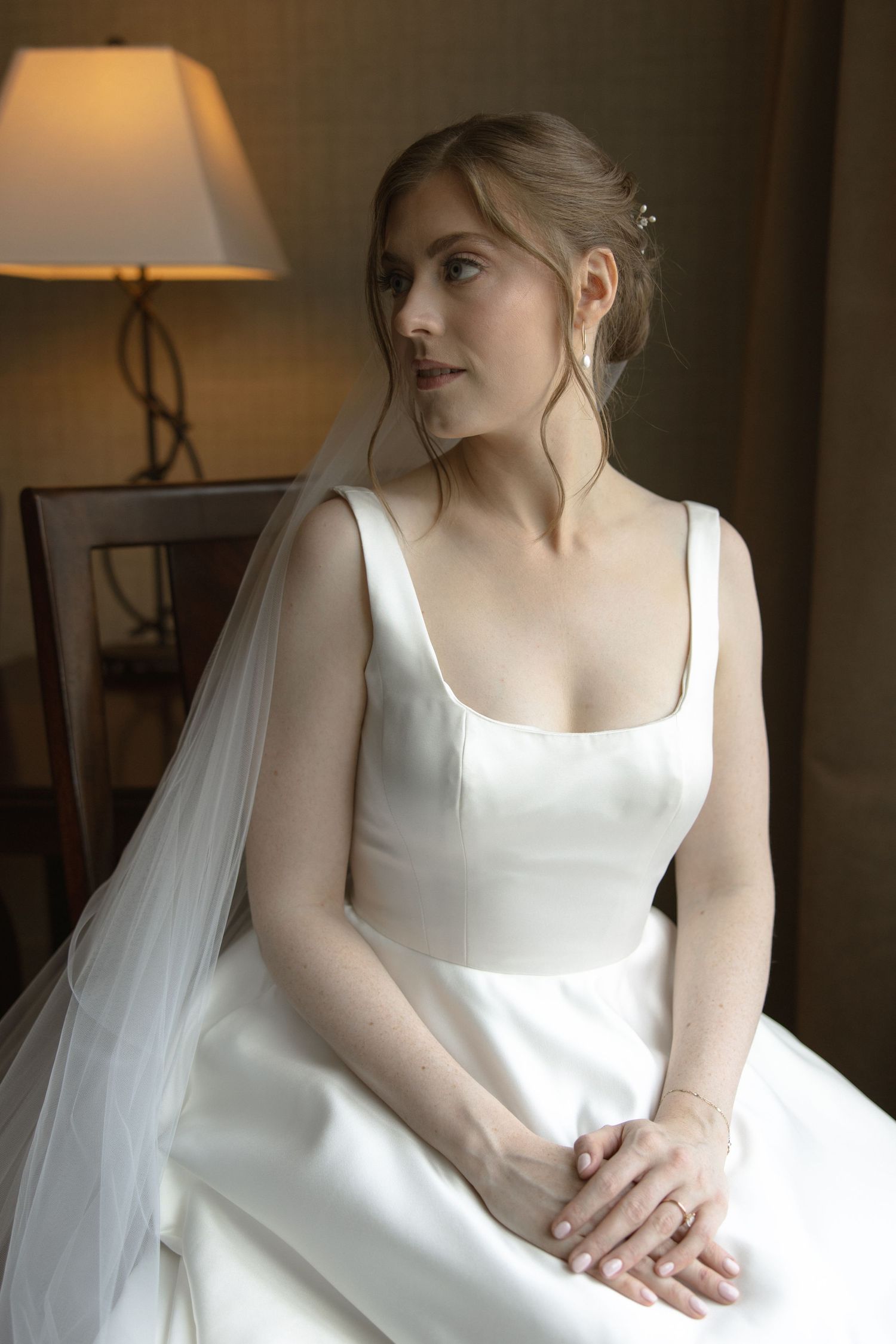 A bride in an elegant white wedding dress with square neckline poses in soft lighting.