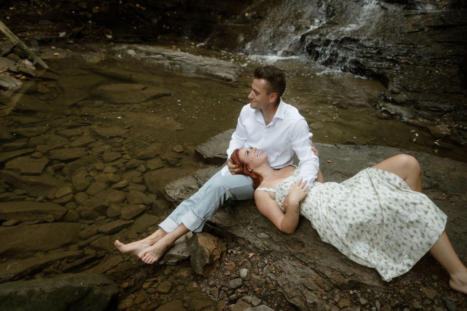 A couple in white attire sits intimately by a rocky creek with a waterfall in the background.