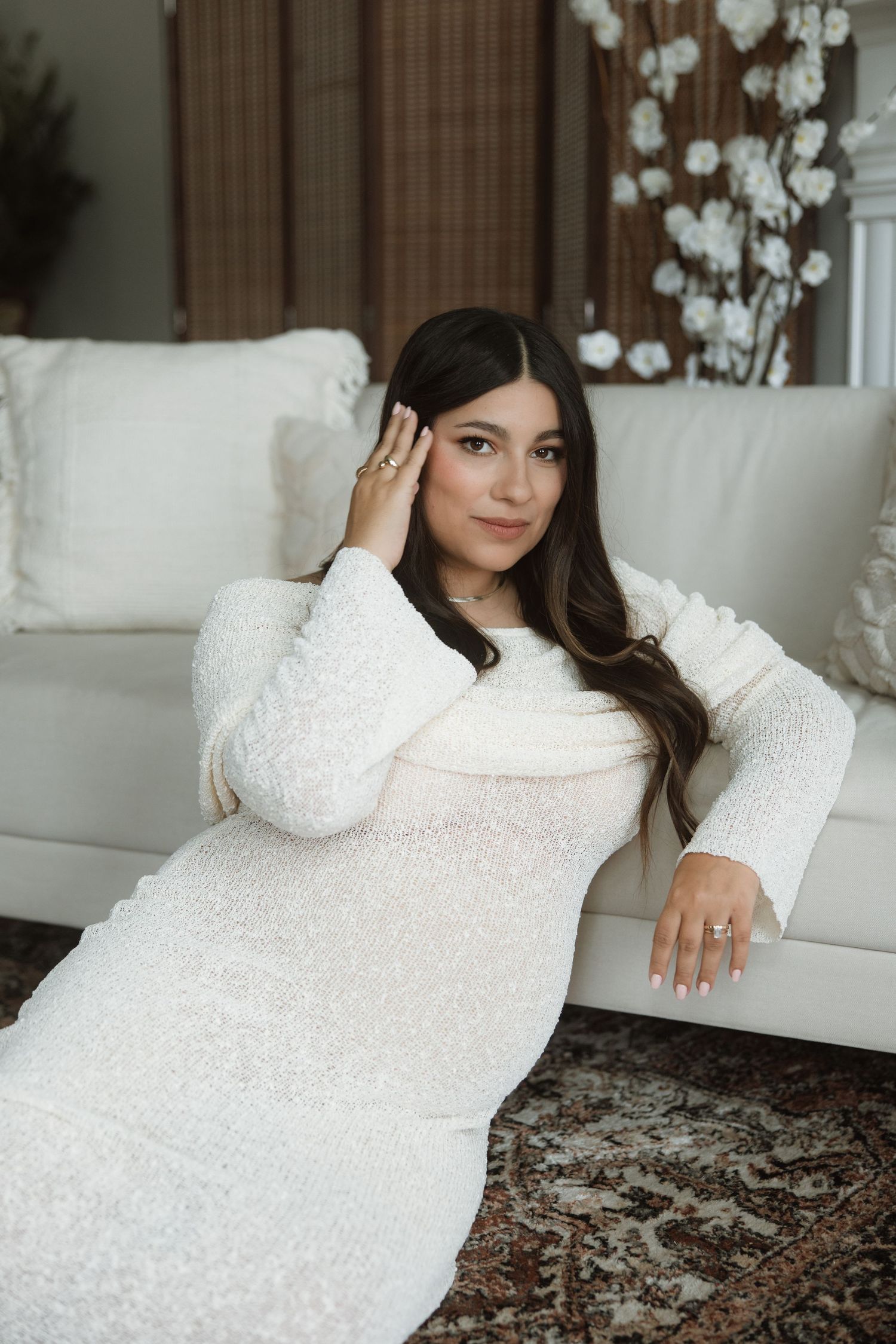 A woman in a white sequined dress poses elegantly on a white sofa with decorative branches in the background.