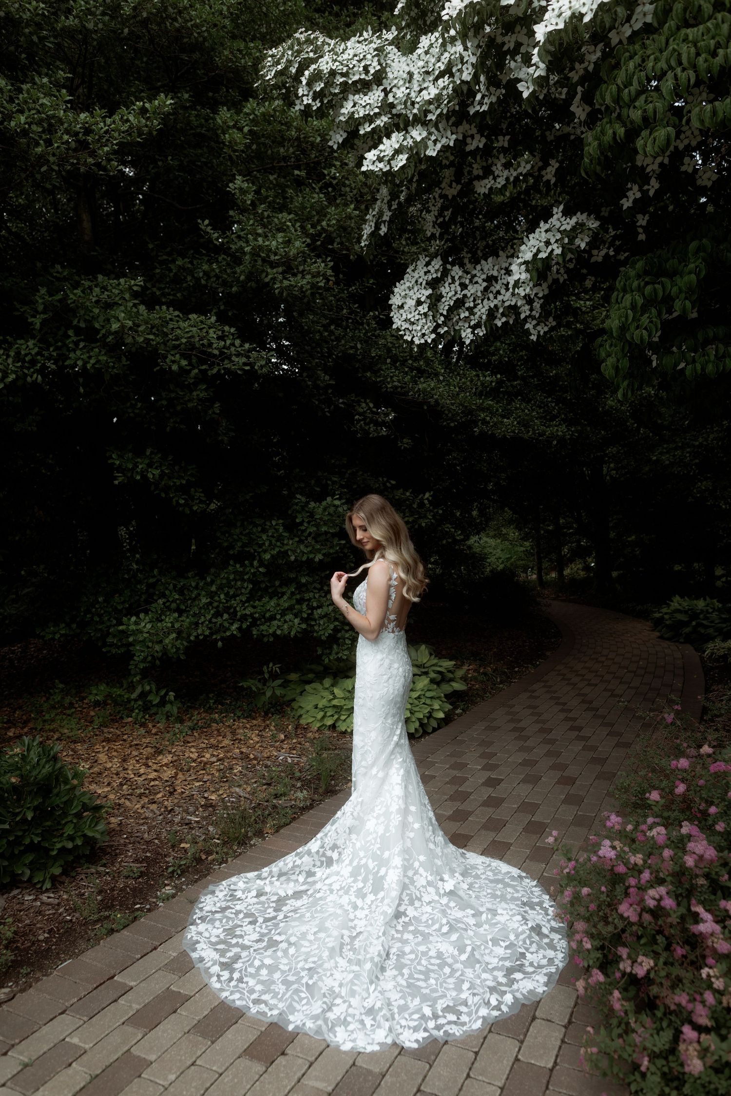 A bride in a lace wedding dress with long train poses on a garden path surrounded by blooming flowers.