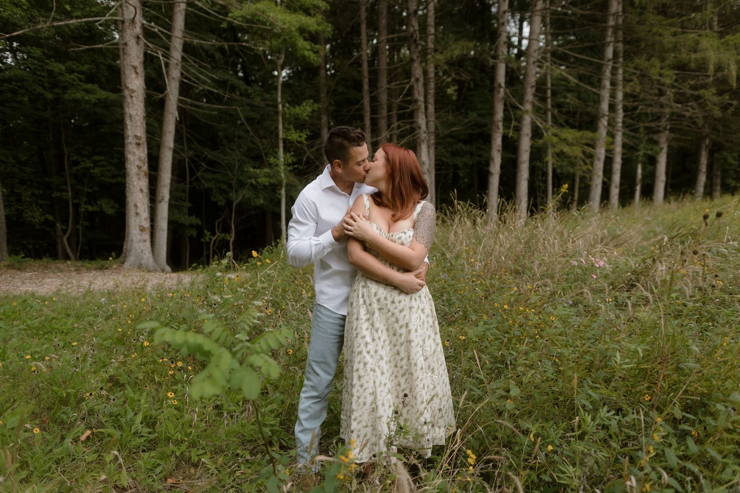 A couple shares a romantic kiss in a sunlit meadow surrounded by tall trees in a forest.