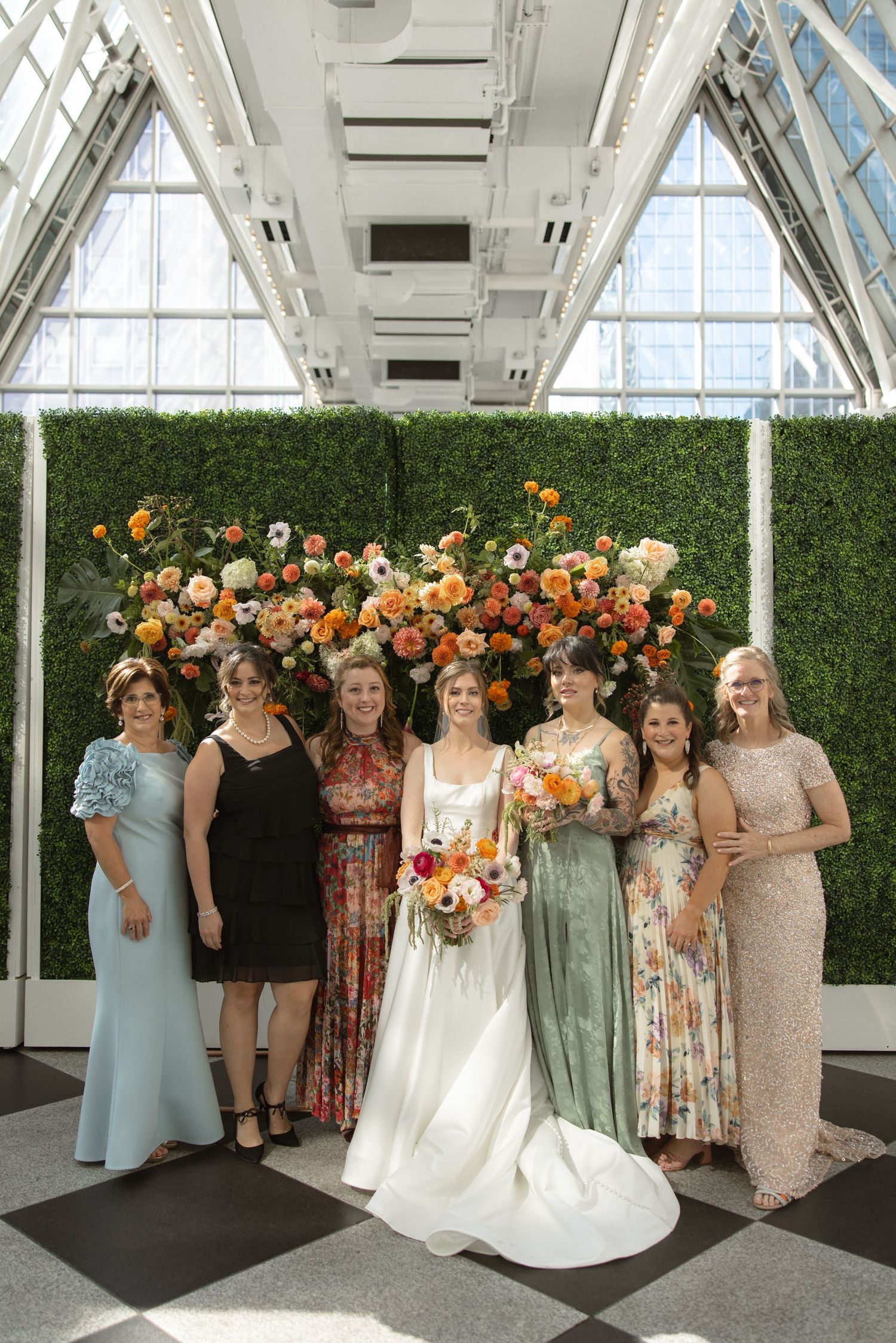 Wedding party in pastel and floral dresses posing against green hedge wall with orange flowers.