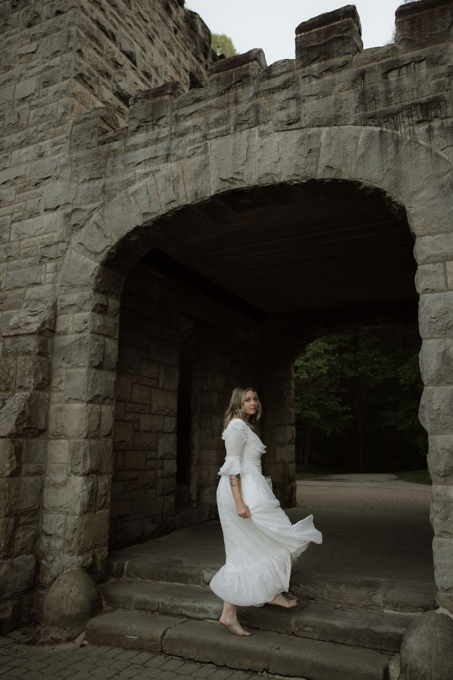 A figure in a flowing white dress stands beneath an ancient stone archway in a moody architectural setting.