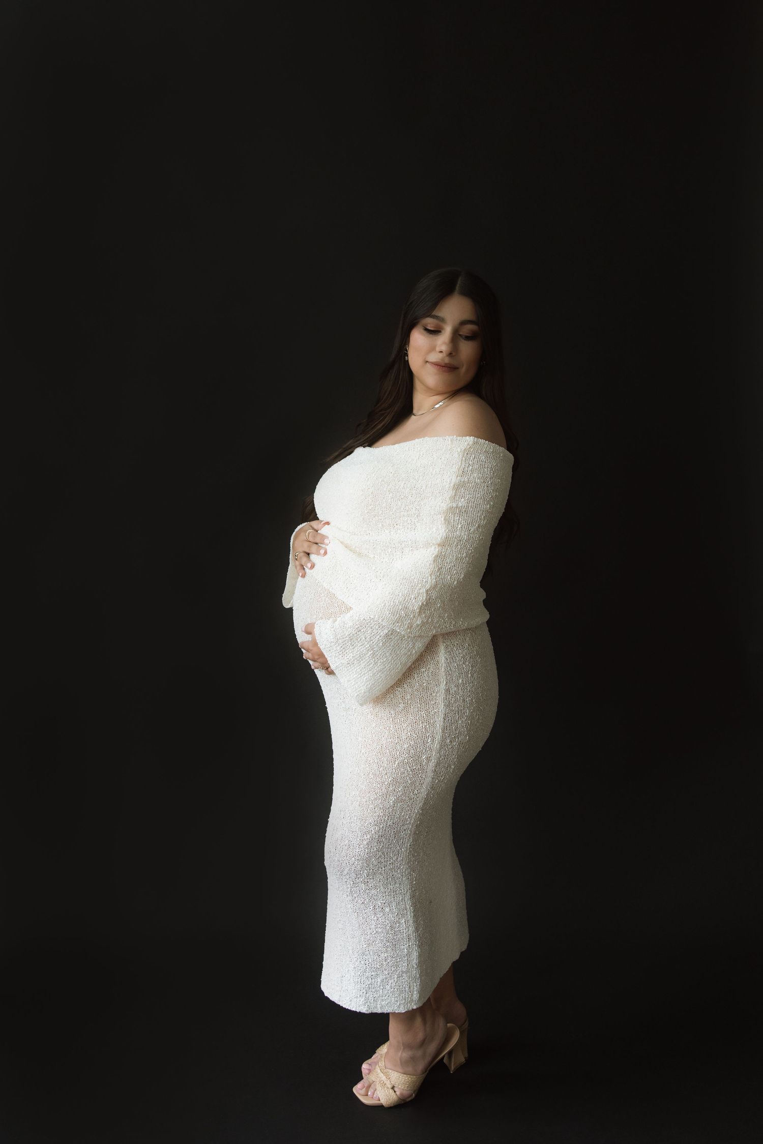 Side view of an elegant maternity portrait with a figure in a fitted white lace dress against a dark background.