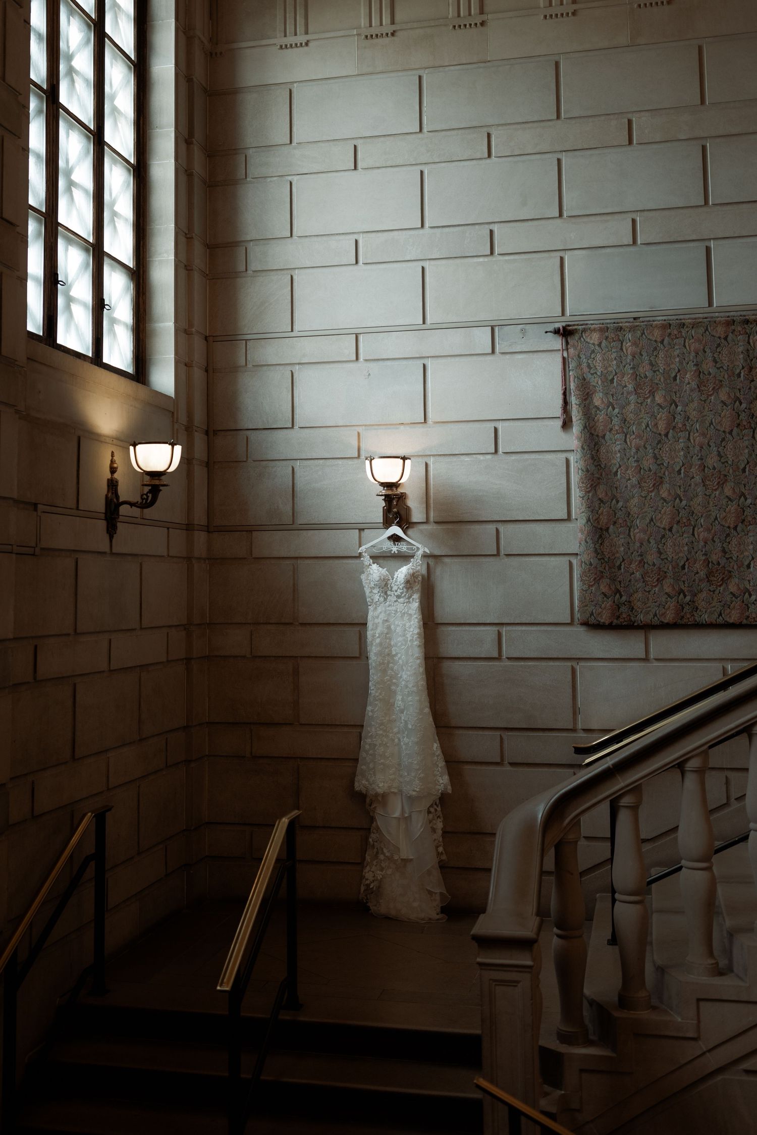 A white lace wedding dress hangs elegantly against a stone wall with sconce lighting near a staircase.