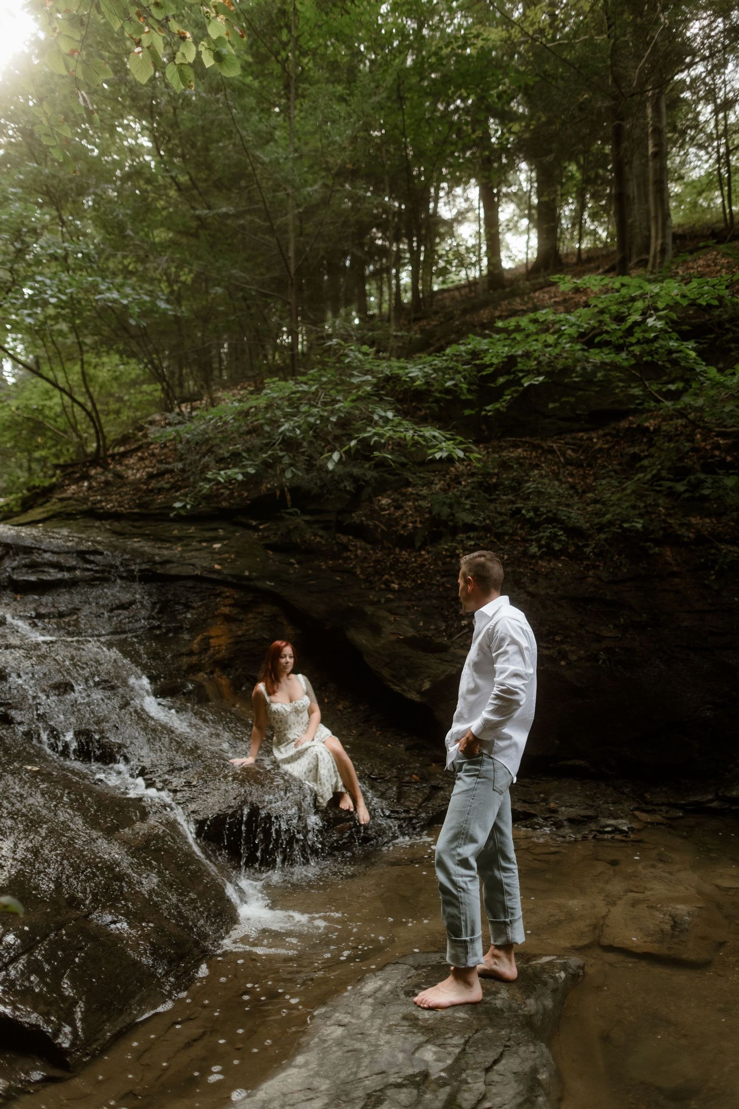 Two people share a romantic moment by a small waterfall in a lush forest setting with sunlight filtering through trees.