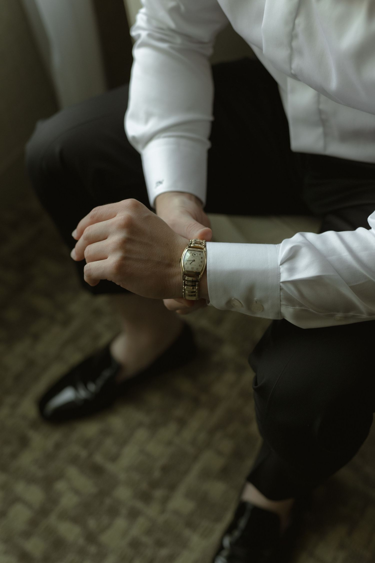 Person in formal attire checking gold watch while sitting on wooden floor.