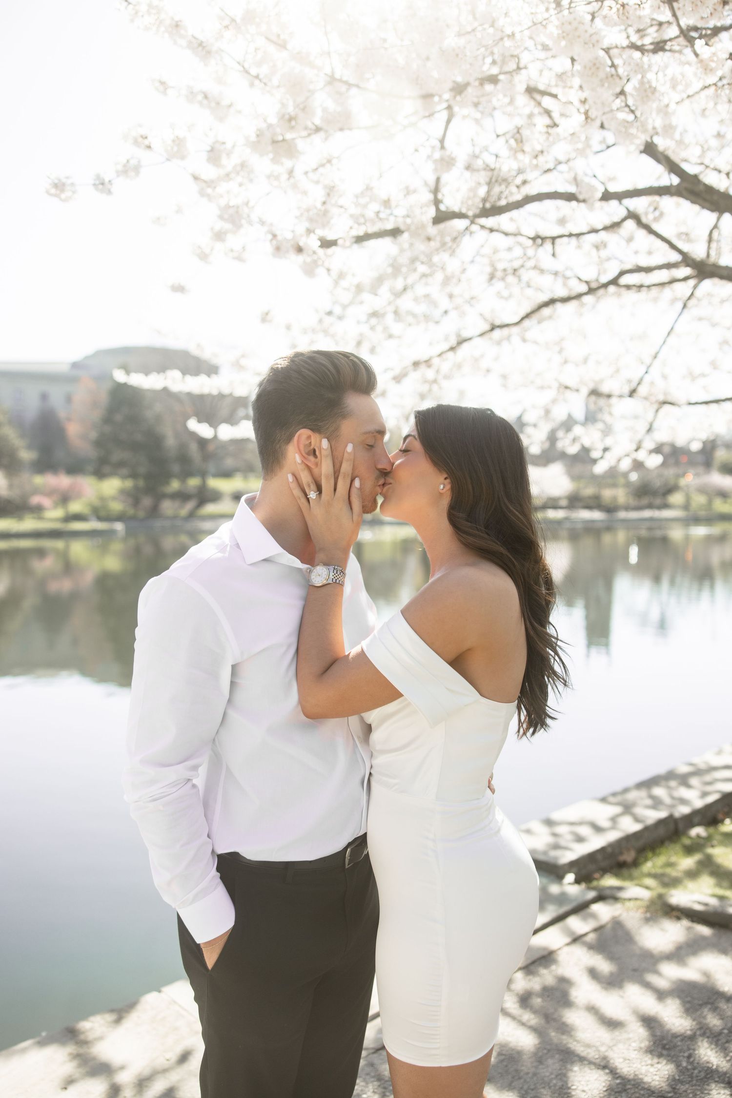 Romantic couple sharing a kiss under blooming cherry blossoms beside a serene lake on a bright spring day.