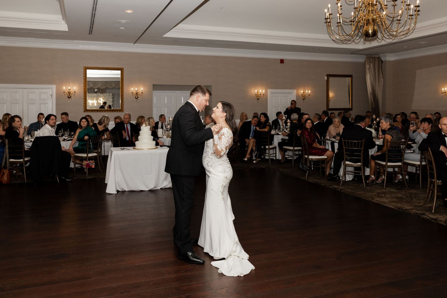 A couple shares their first dance at their wedding reception in an elegant ballroom with chandeliers and round tables.