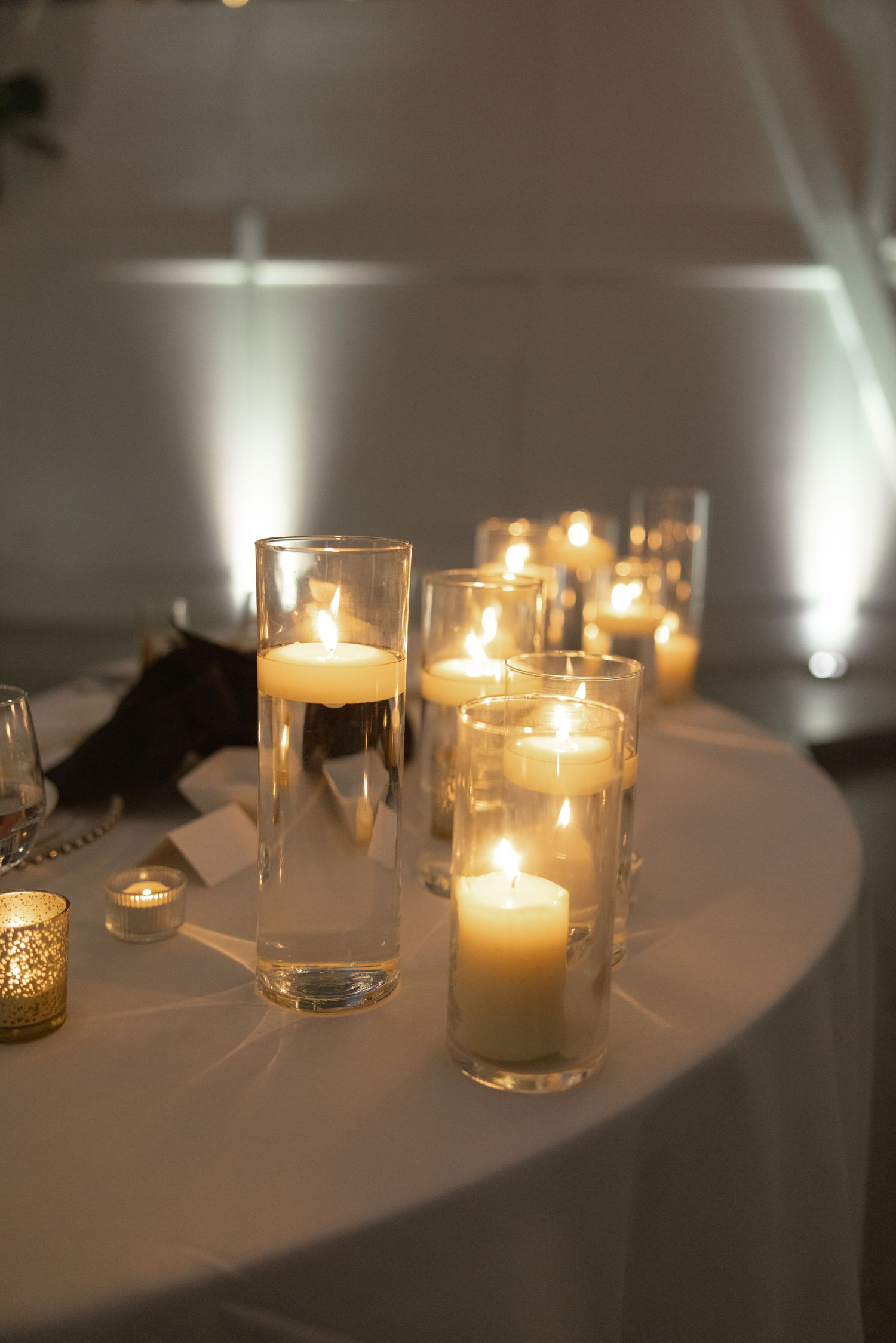 Elegant wedding reception table setting with glowing candles in glass cylinders on white tablecloth.