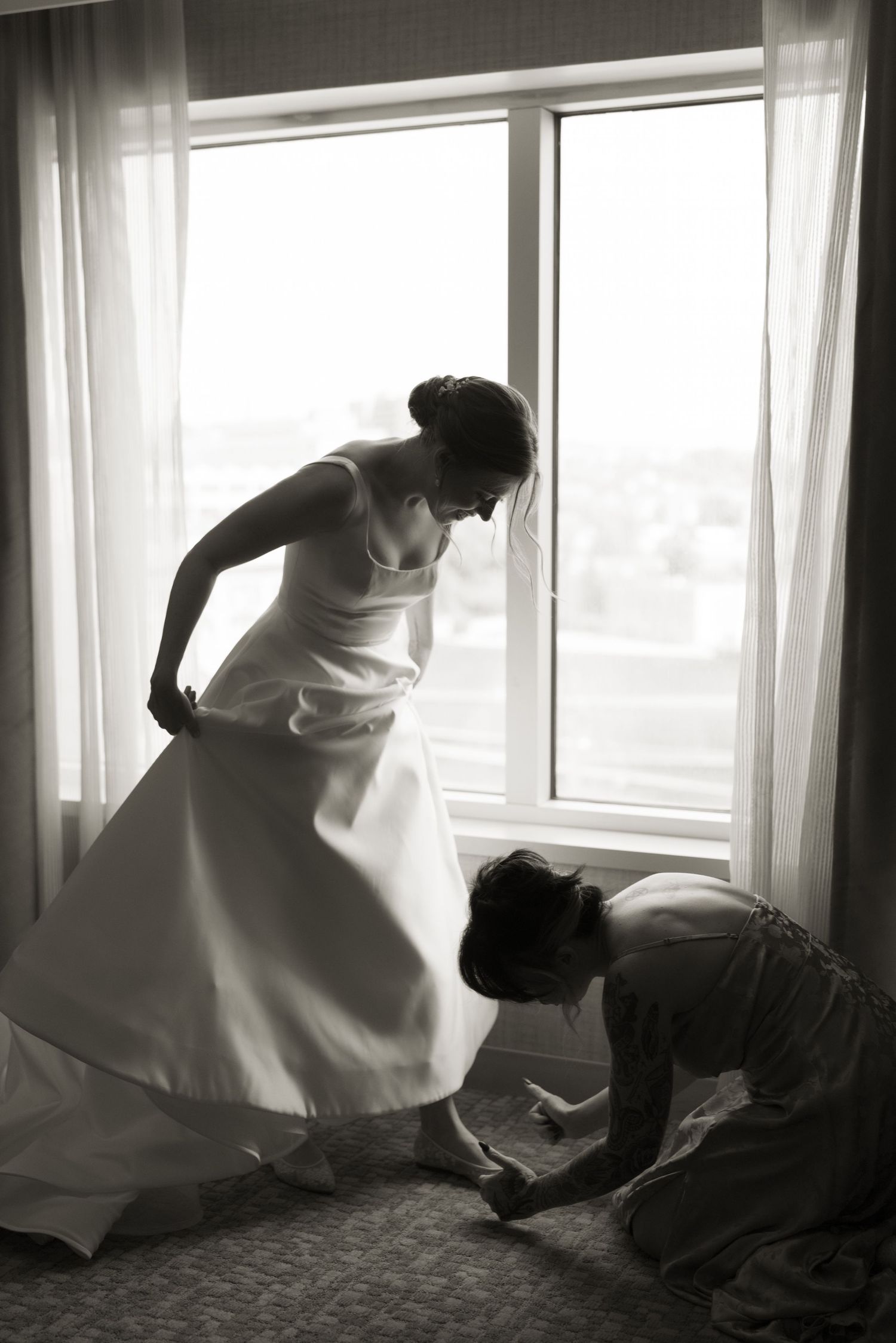 Black and white image of wedding dress preparations with dramatic window lighting.