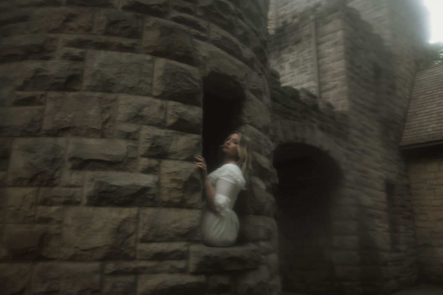A ghostly figure in white clothing stands against a dark stone castle wall in a blurred, atmospheric photo.