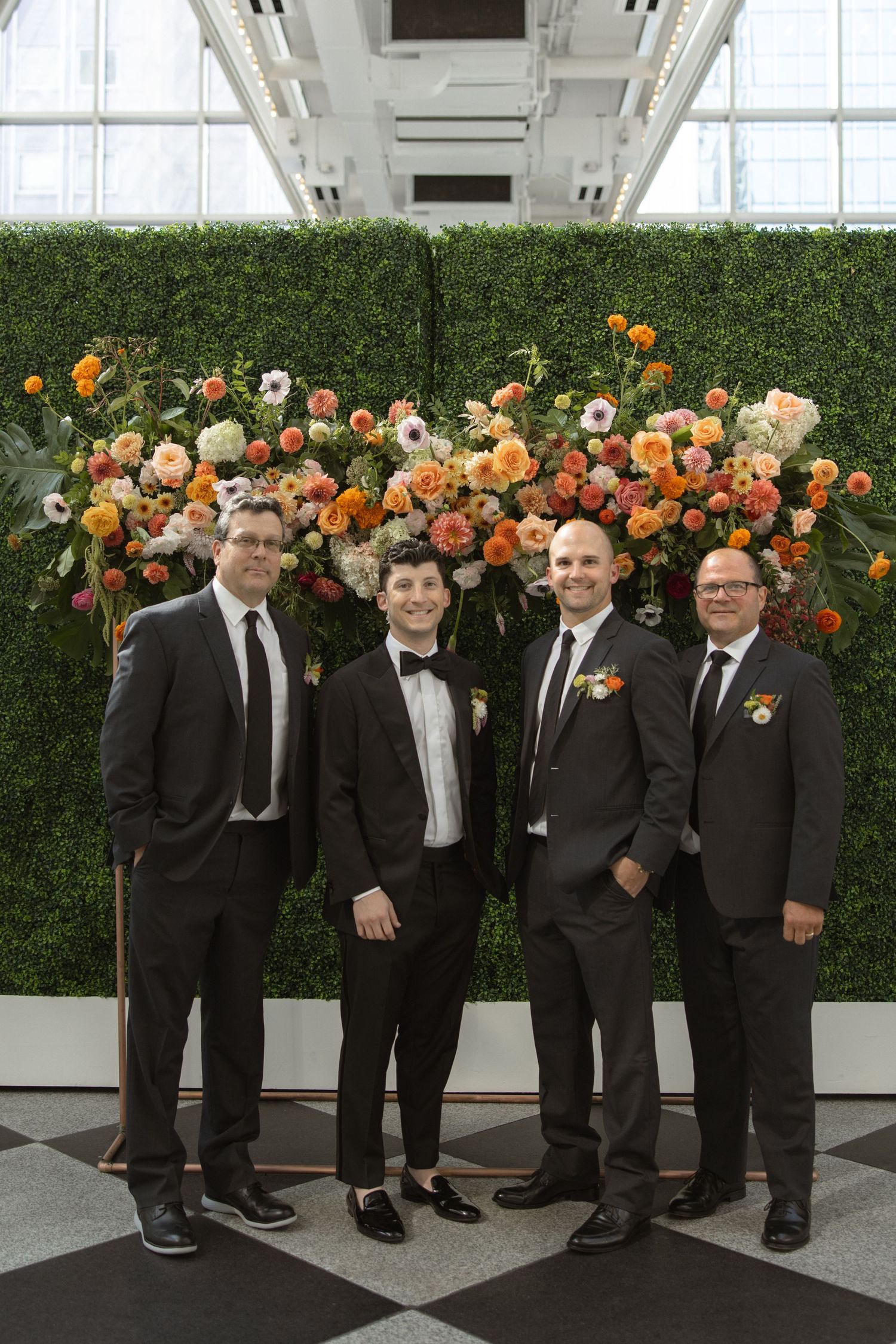 Groomsmen in dark suits pose against floral wall with orange and cream blooms.