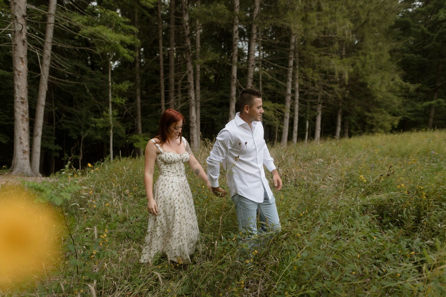 Two people in a romantic outdoor setting walk together through tall grass with pine trees in the background.